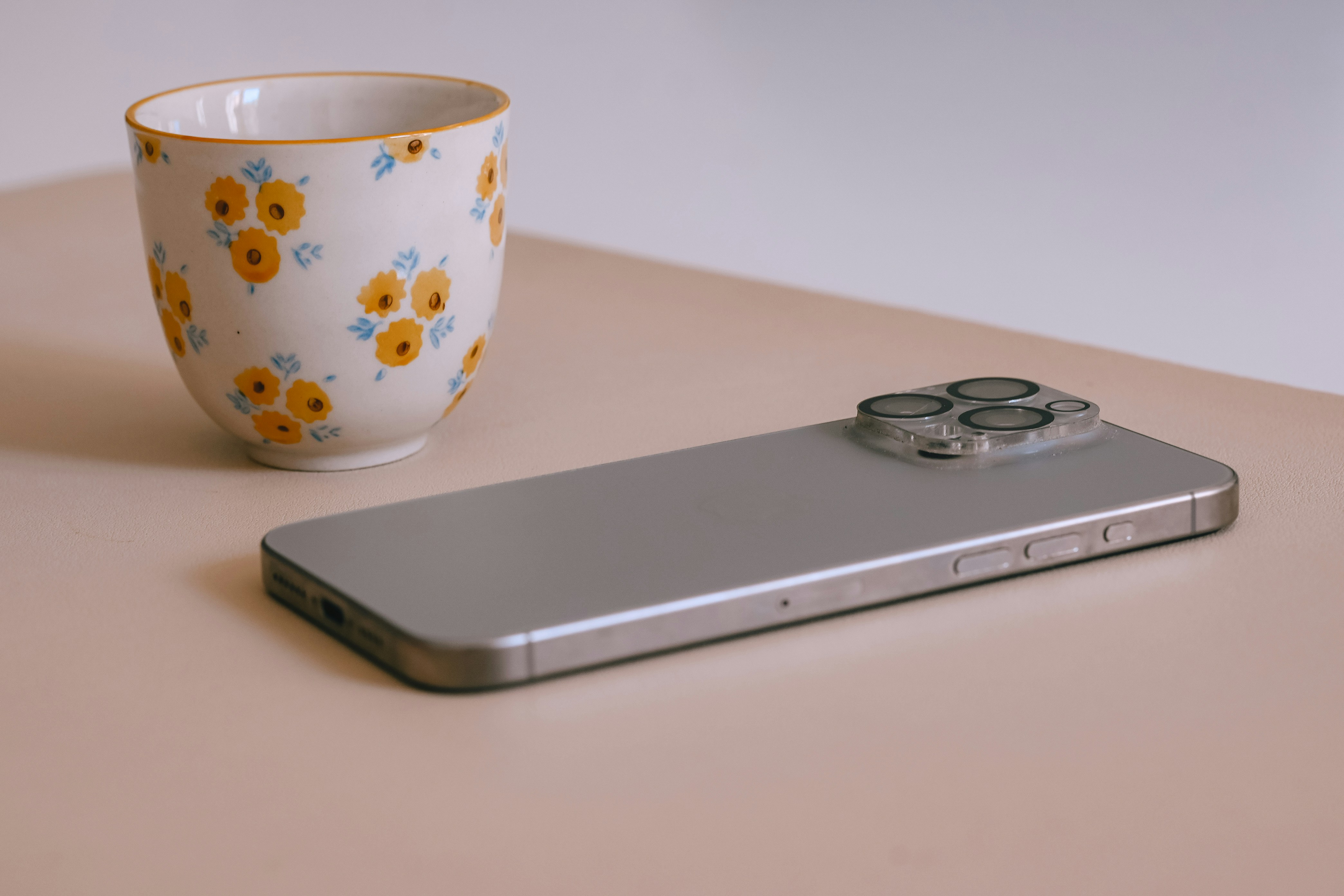A silver smartphone lies next to a floral patterned cup.