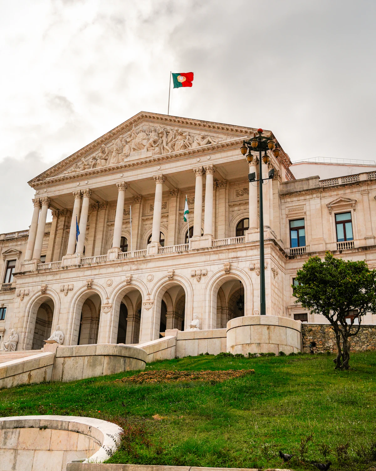 Exterior da Assembleia da República portuguesa, com a bandeira nacional sob um céu nublado.