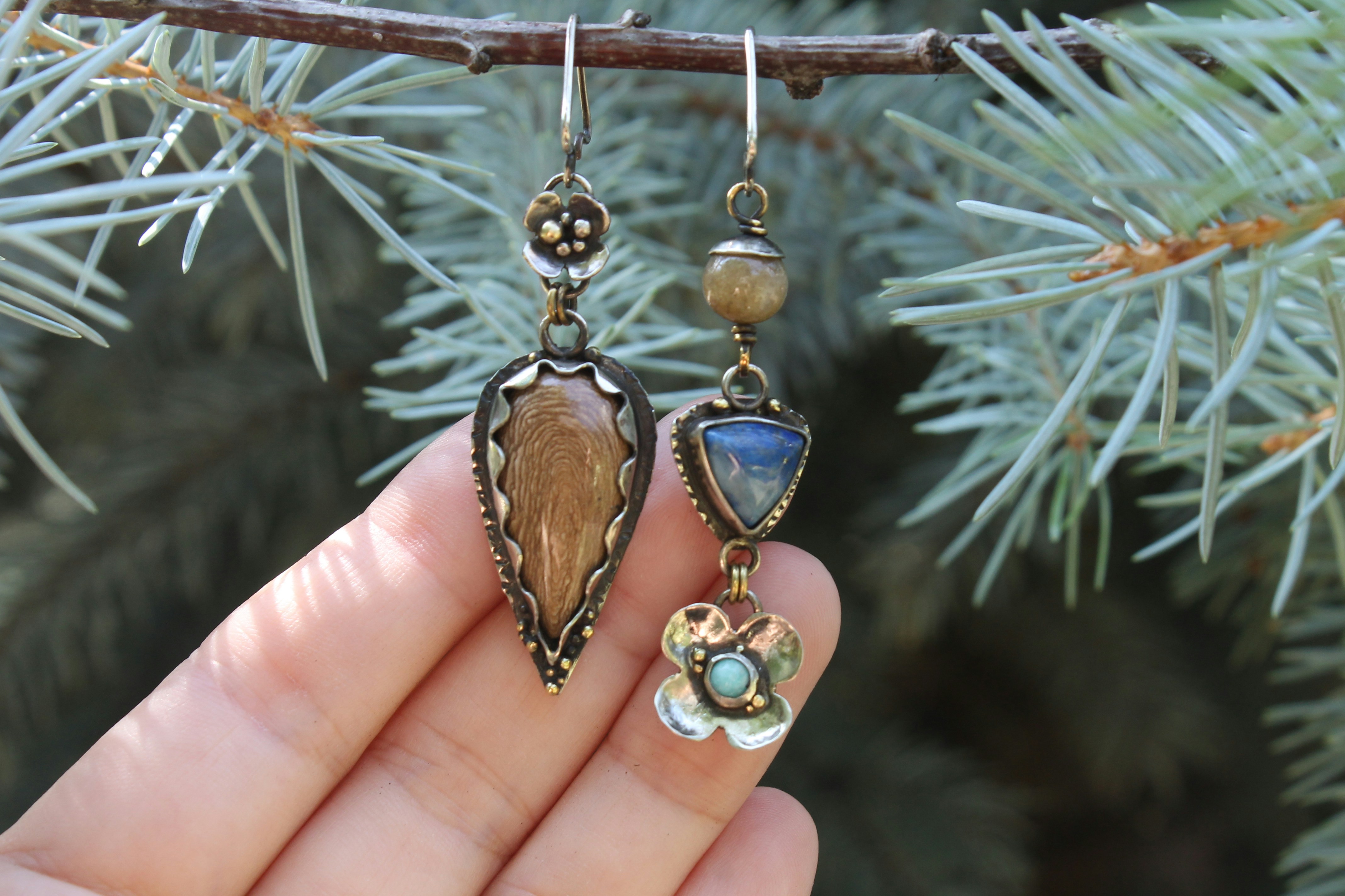 Une main tient deux boucles d’oreilles uniques contre une branche de pin