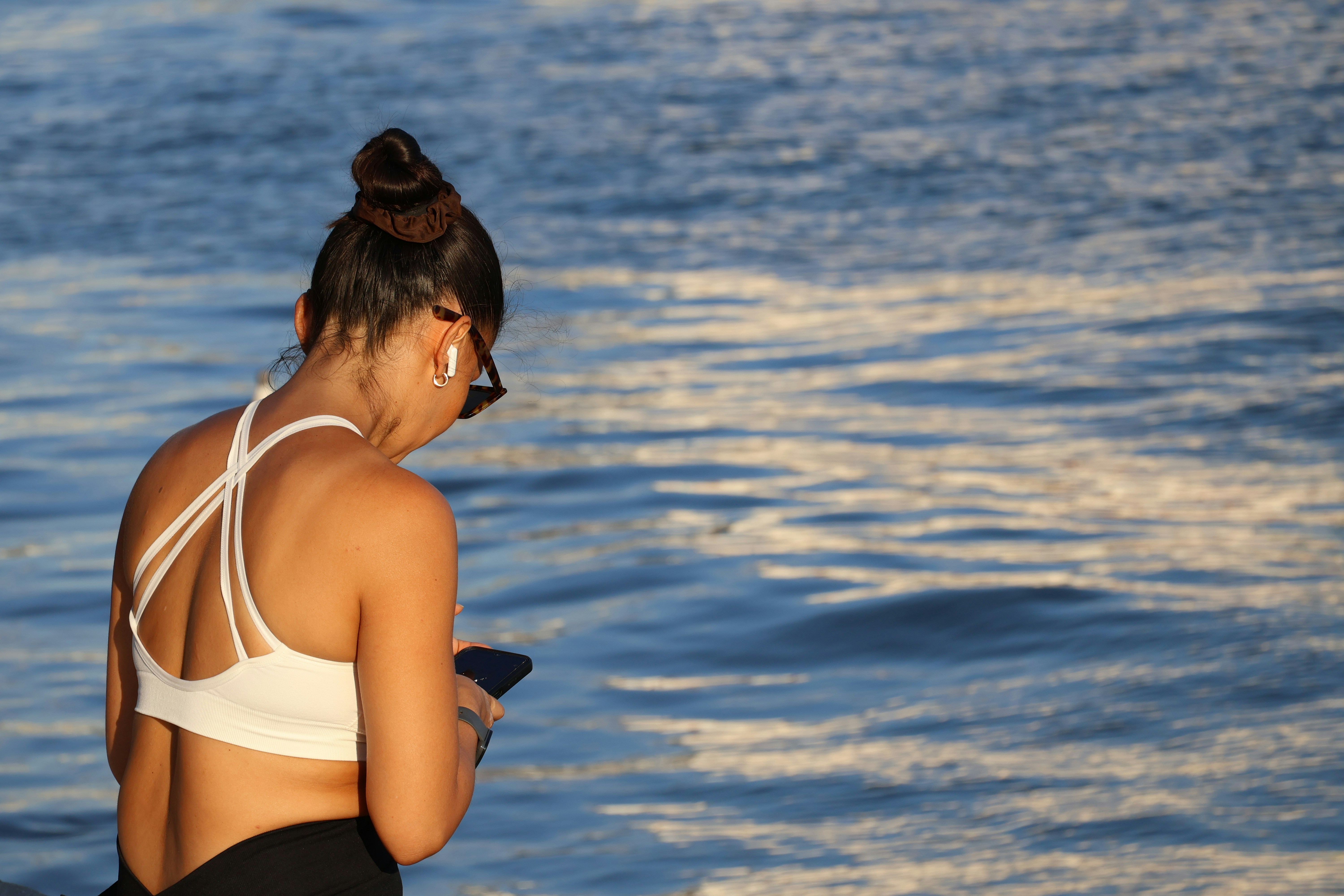 スポーツブラ姿の女性が水辺でスマホを見ている。