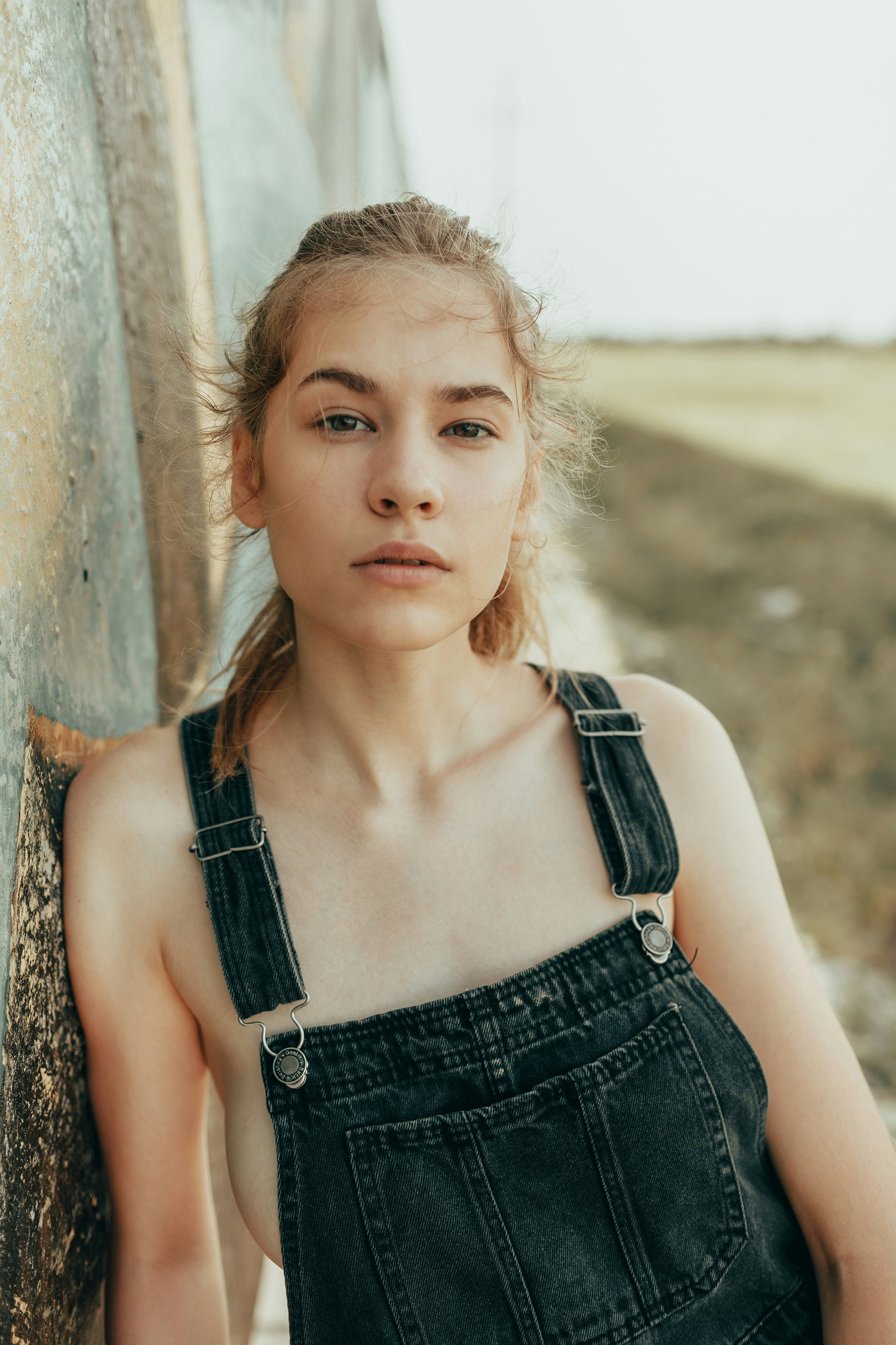 A young woman in denim overalls leaning against a wall.