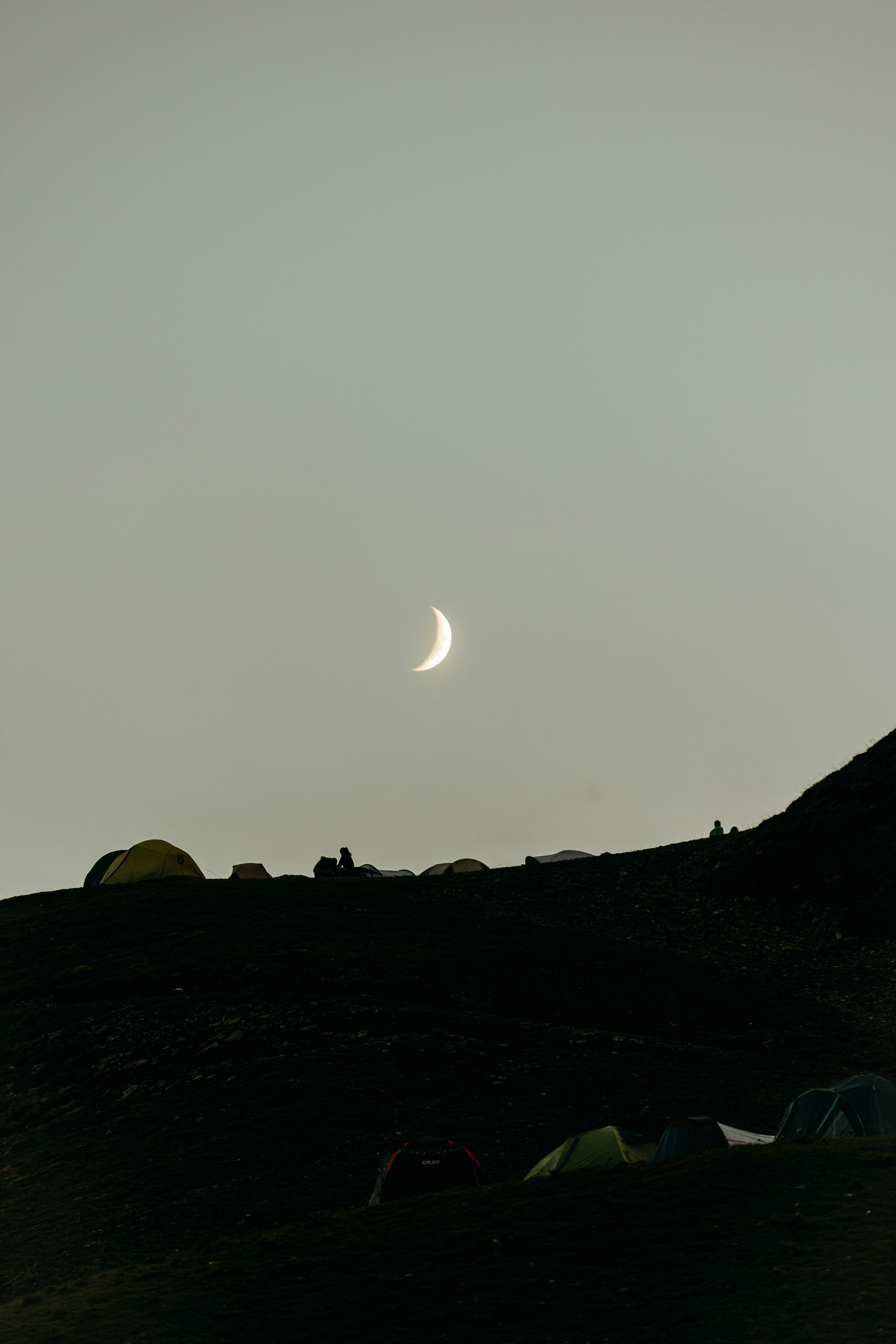 A crescent moon hangs over dark hills with tents.