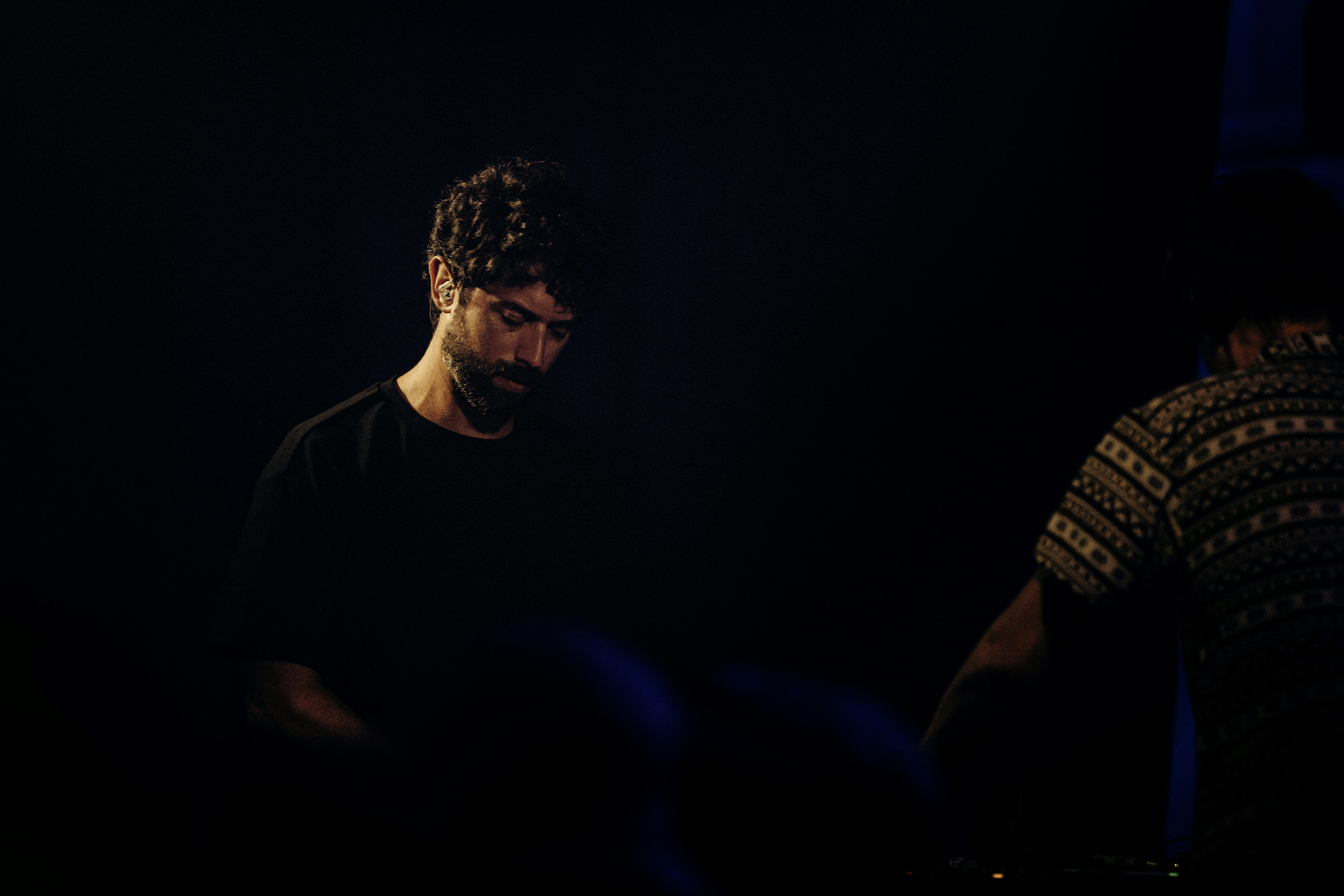 Bearded man in a dark room, another person nearby.