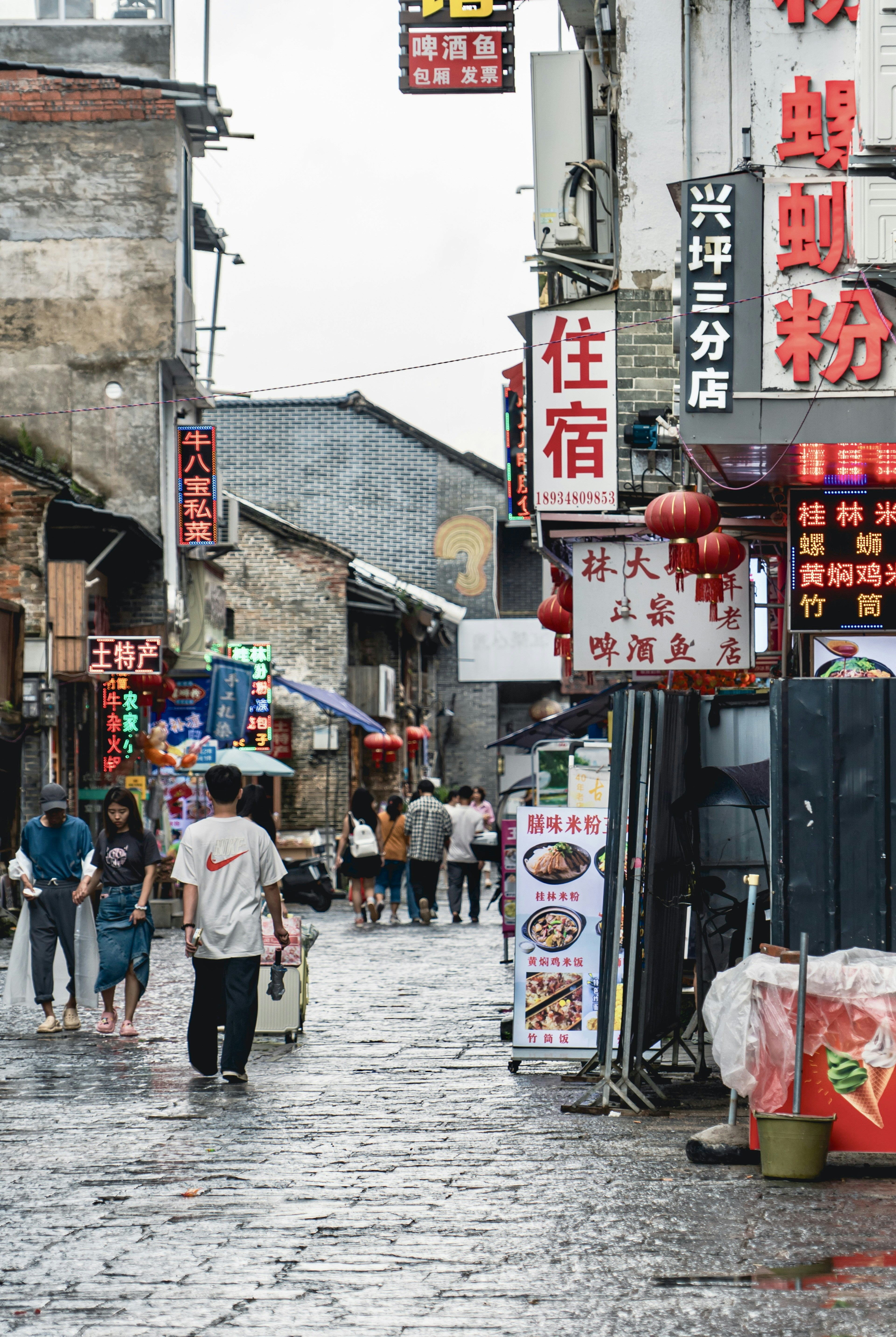 Pedestrians walk down a narrow, wet street with glowing signs.
