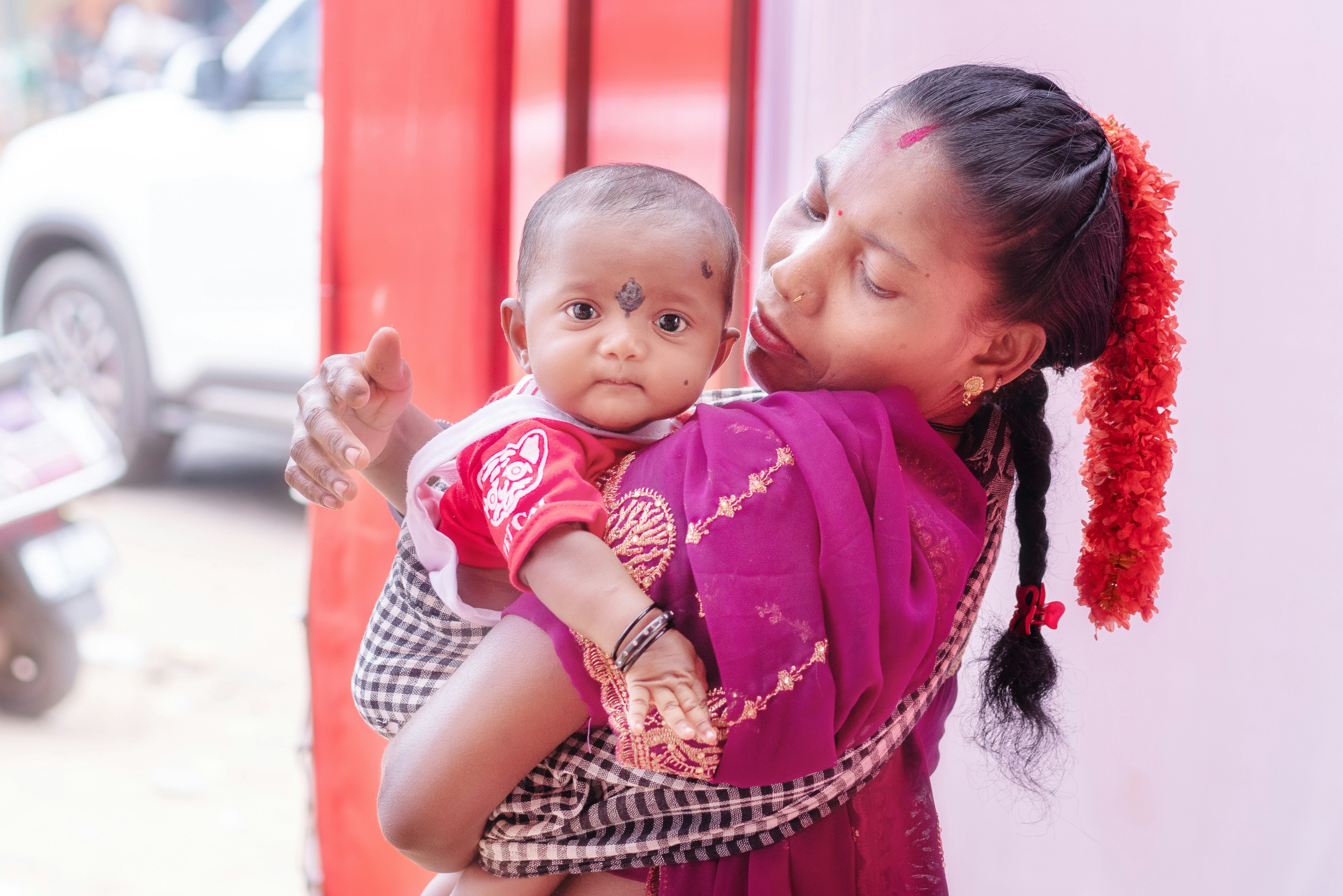 Woman holds baby, adorned with red flowers and bindi.