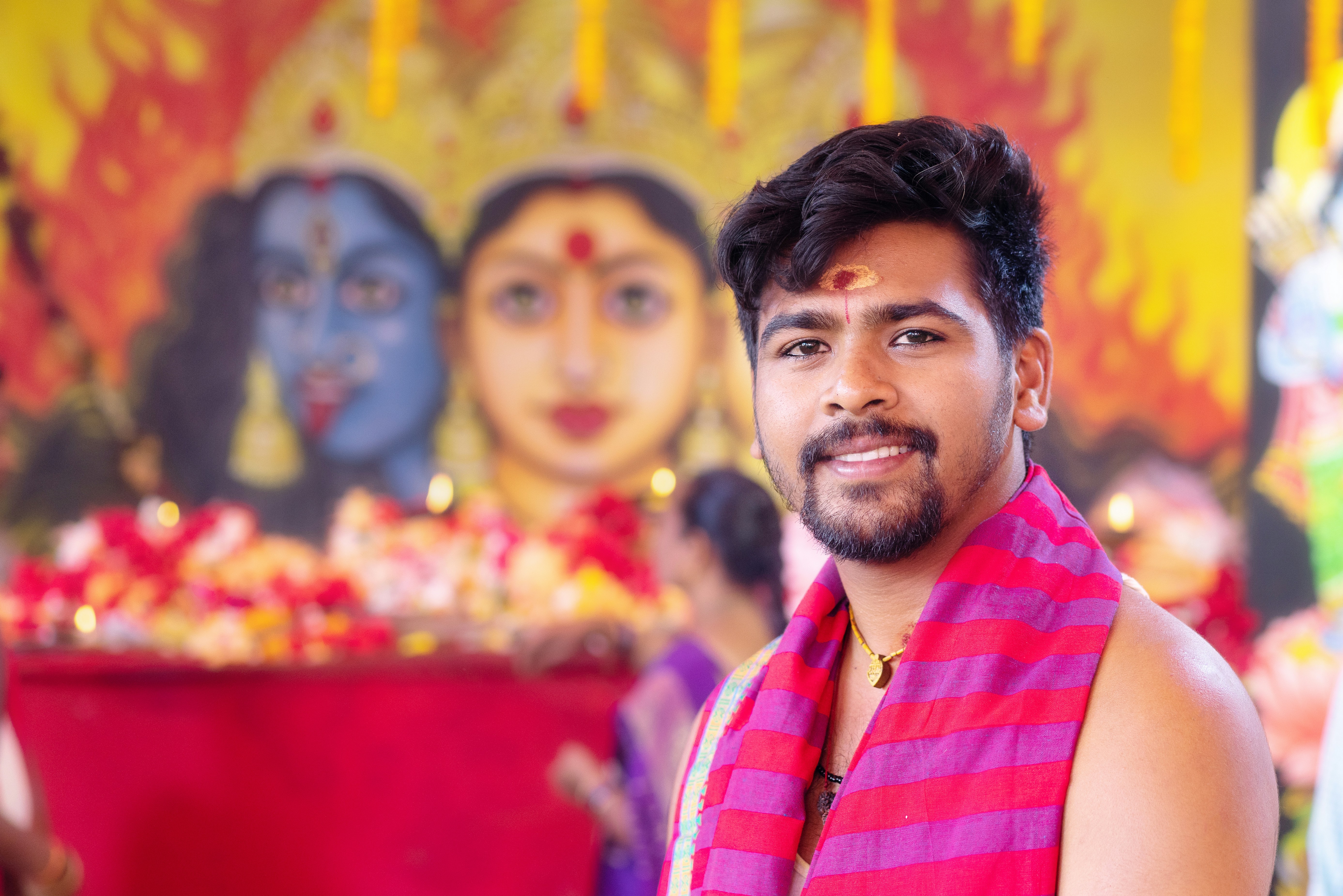 Smiling man with tilak in front of deity paintings.