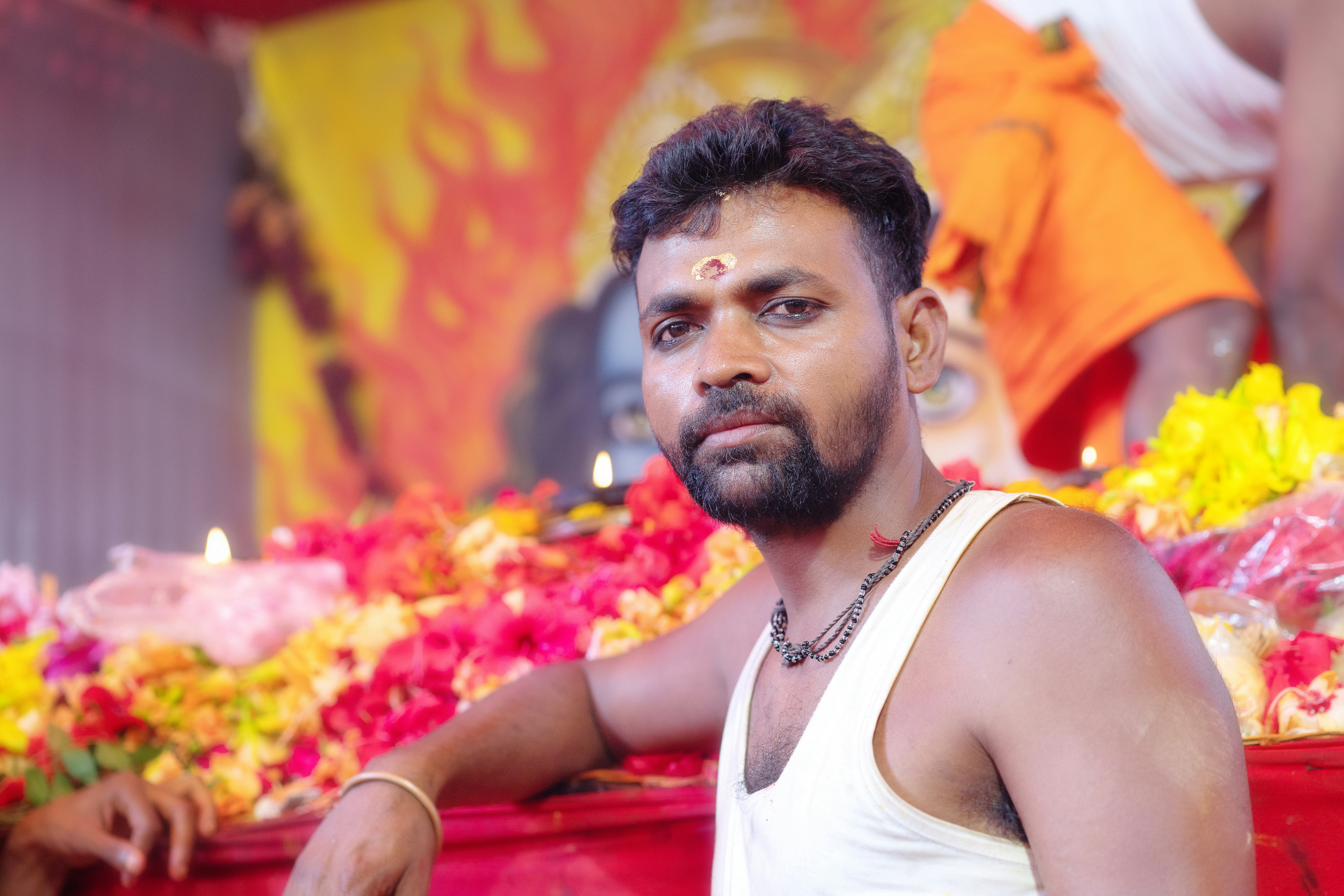 Bearded man with bindi among colorful flowers at a ceremony.