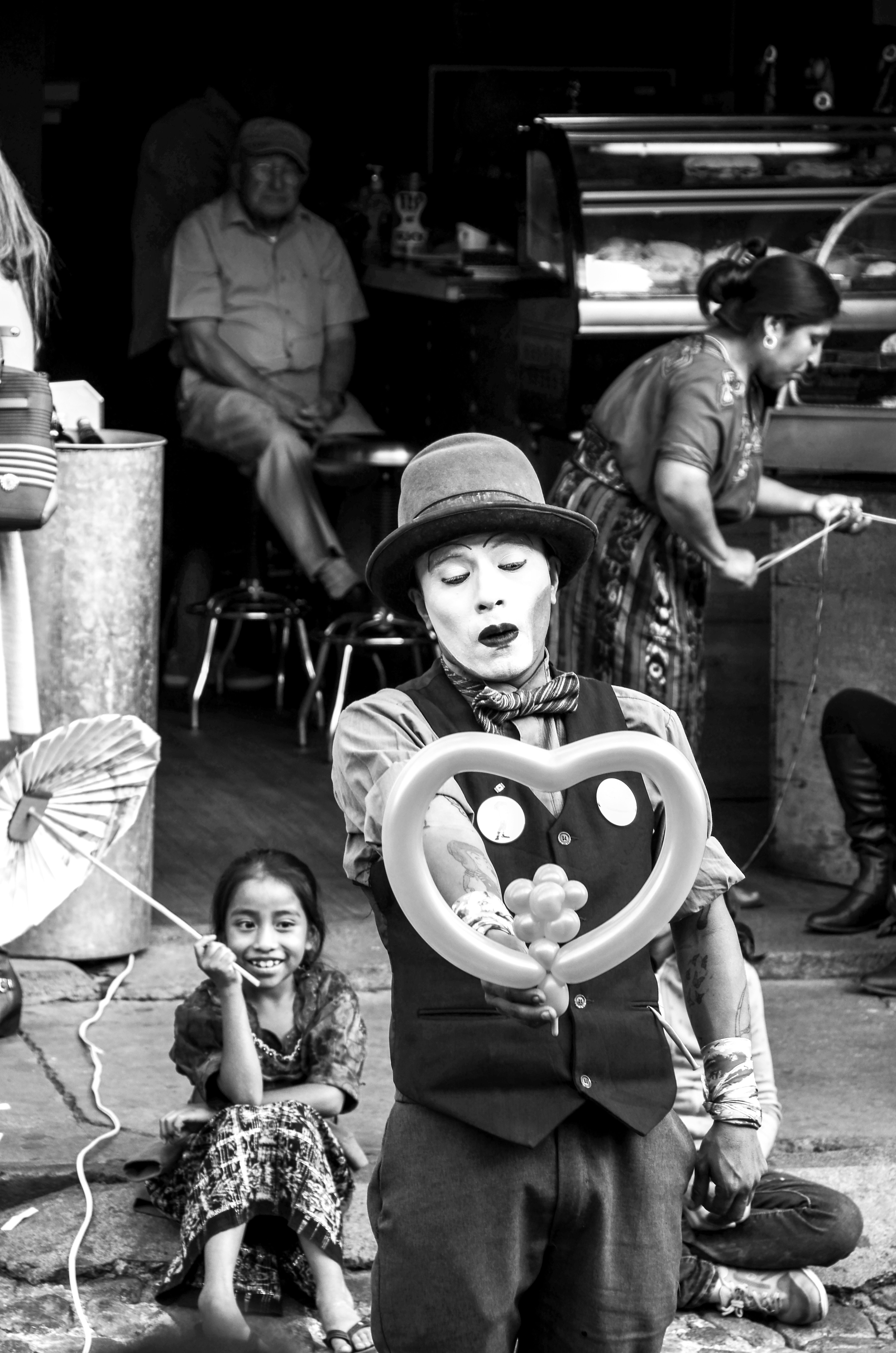 Clown holds heart balloon, girl smiles, black and white street.