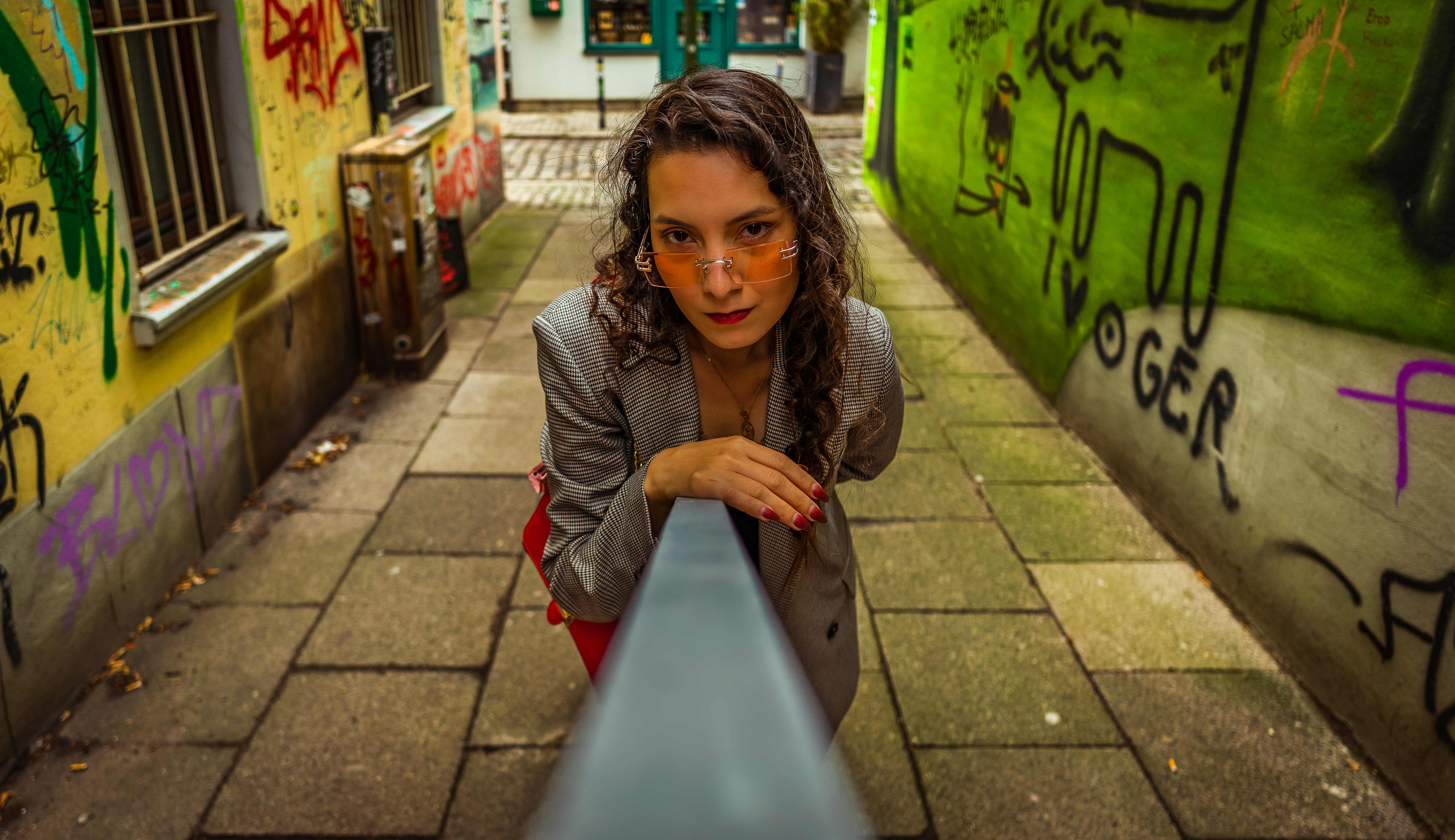 Young woman in blazer leans on railing in graffiti alley.