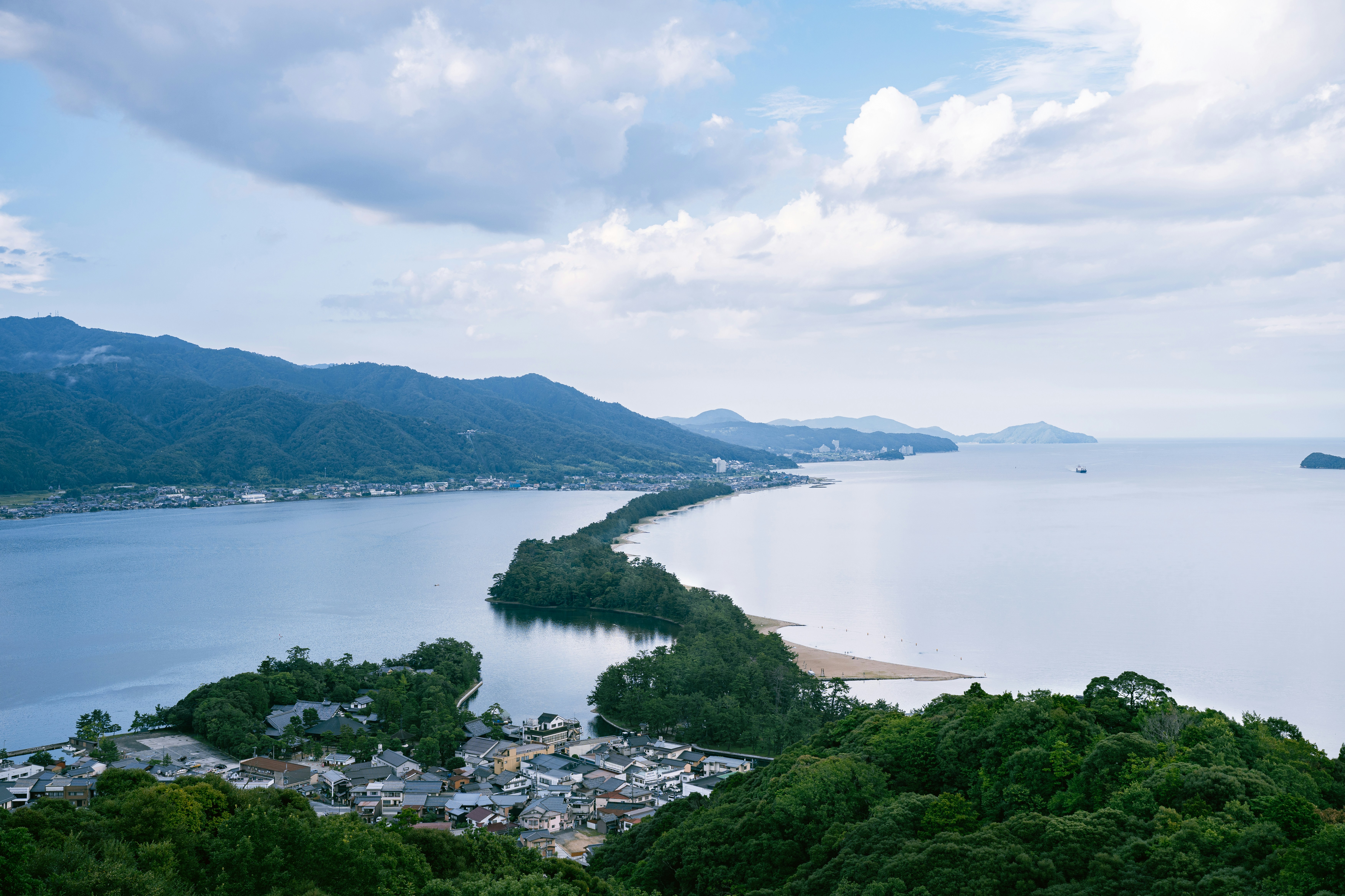 Long, tree-covered sandspit separates water, with mountains and a town