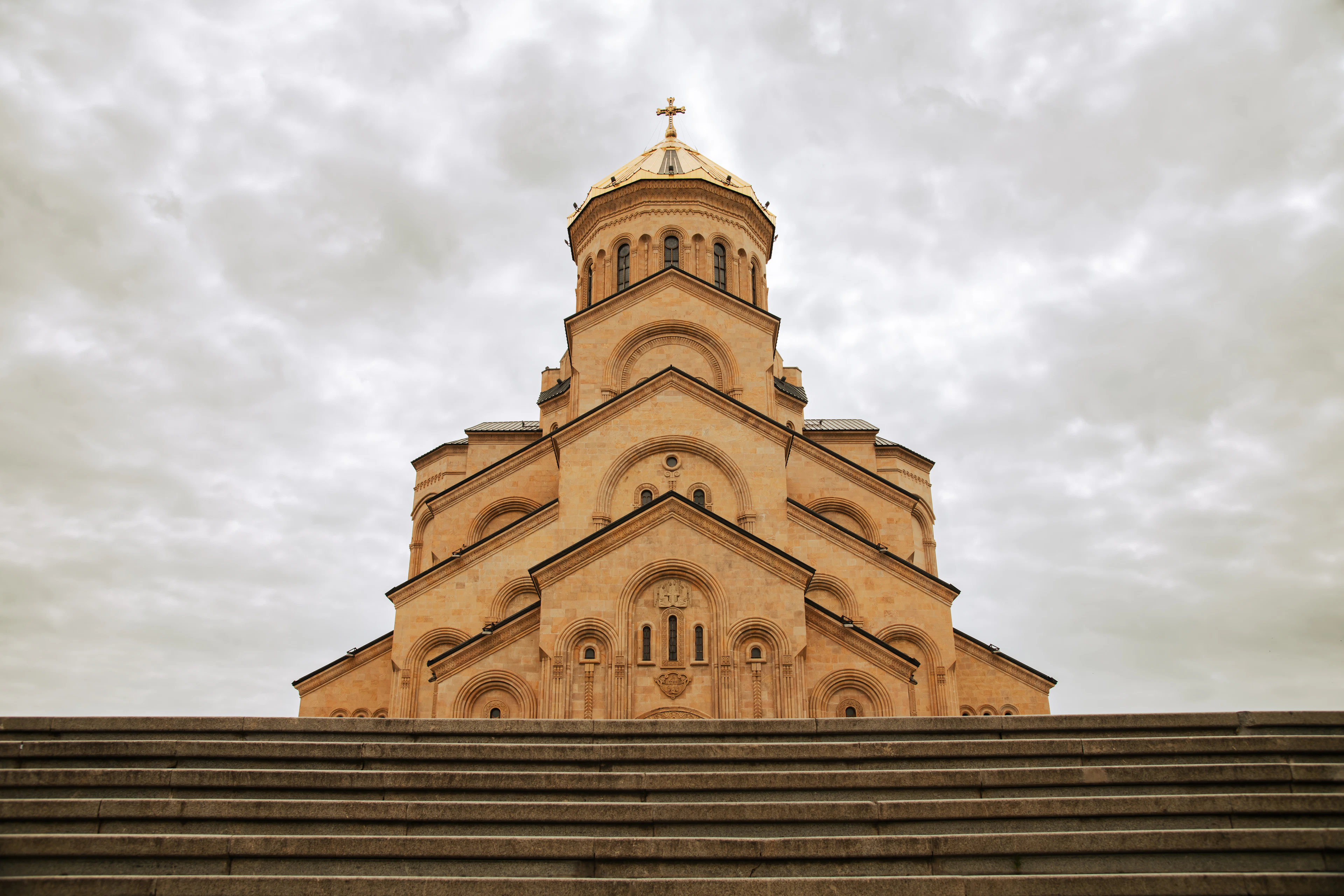 The Holy Trinity Cathedral of Tbilisi seen head-on from the foot of its steps, tier upon tier of pale sandstone rising to a gilded dome and cross, silhouetted against a bank of heavy grey cloud