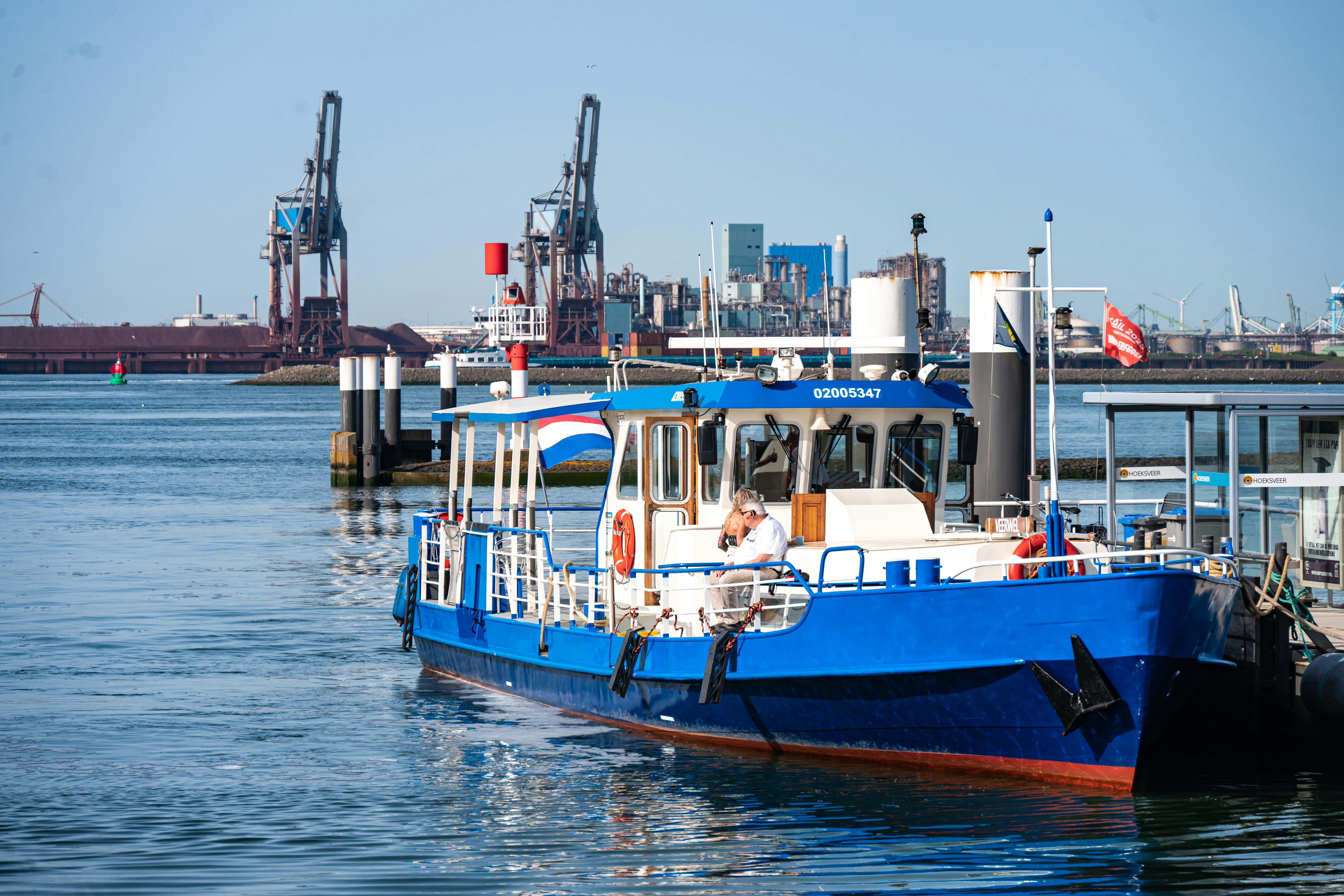 Blue passenger boat docked in a sunny industrial port.