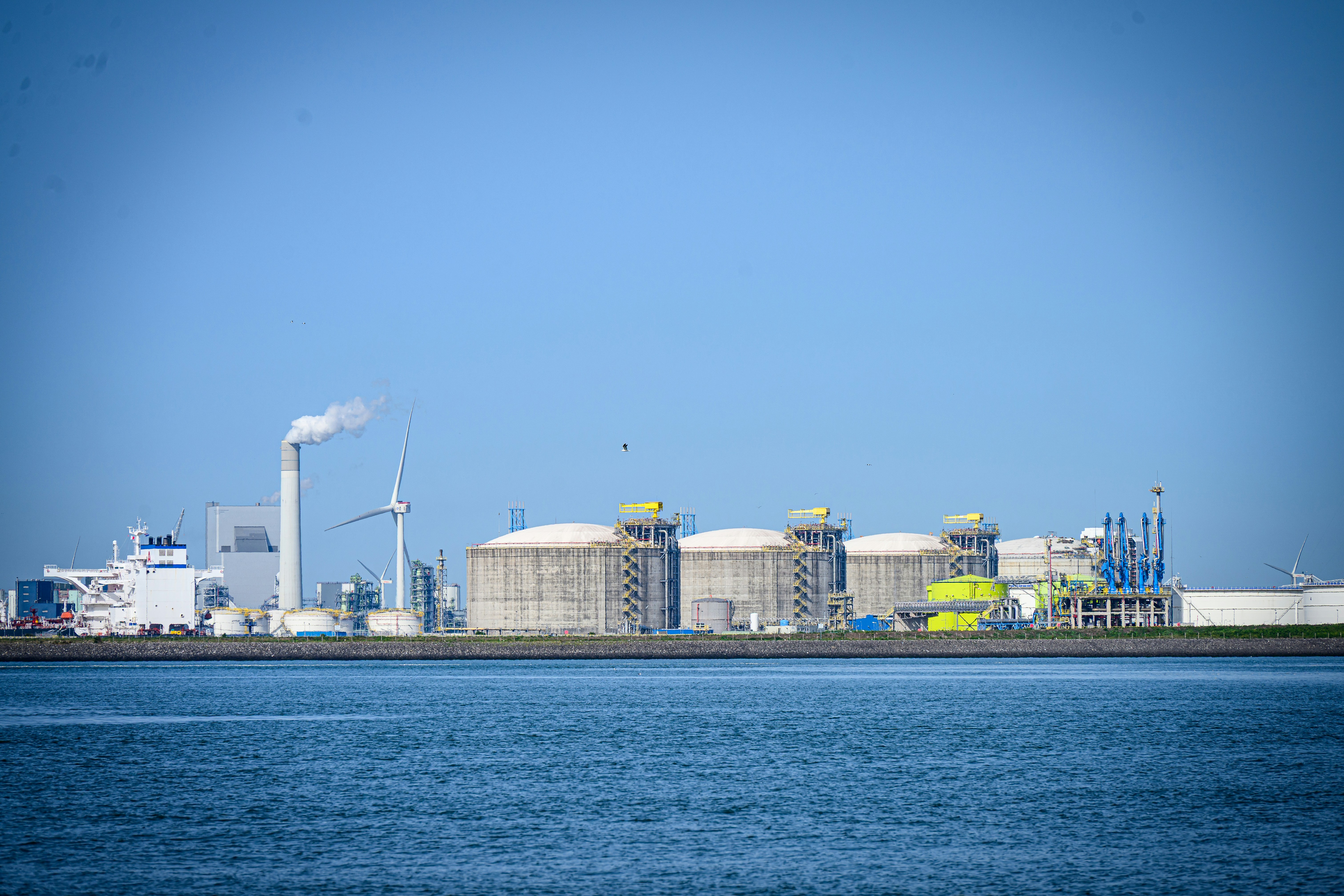 Industrial complex with tanks, factory, and wind turbines by water.