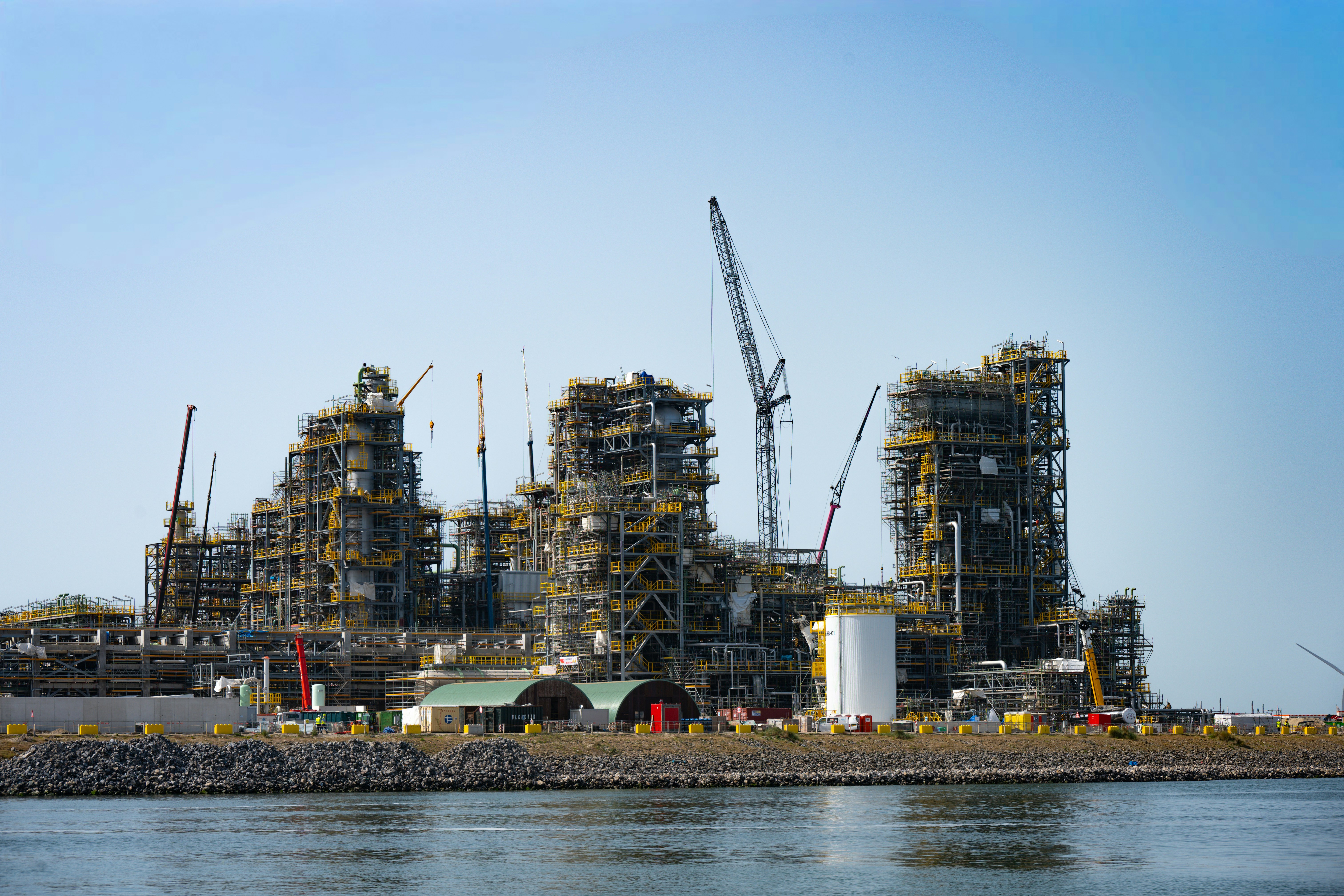 Large industrial plant under construction near water with cranes.