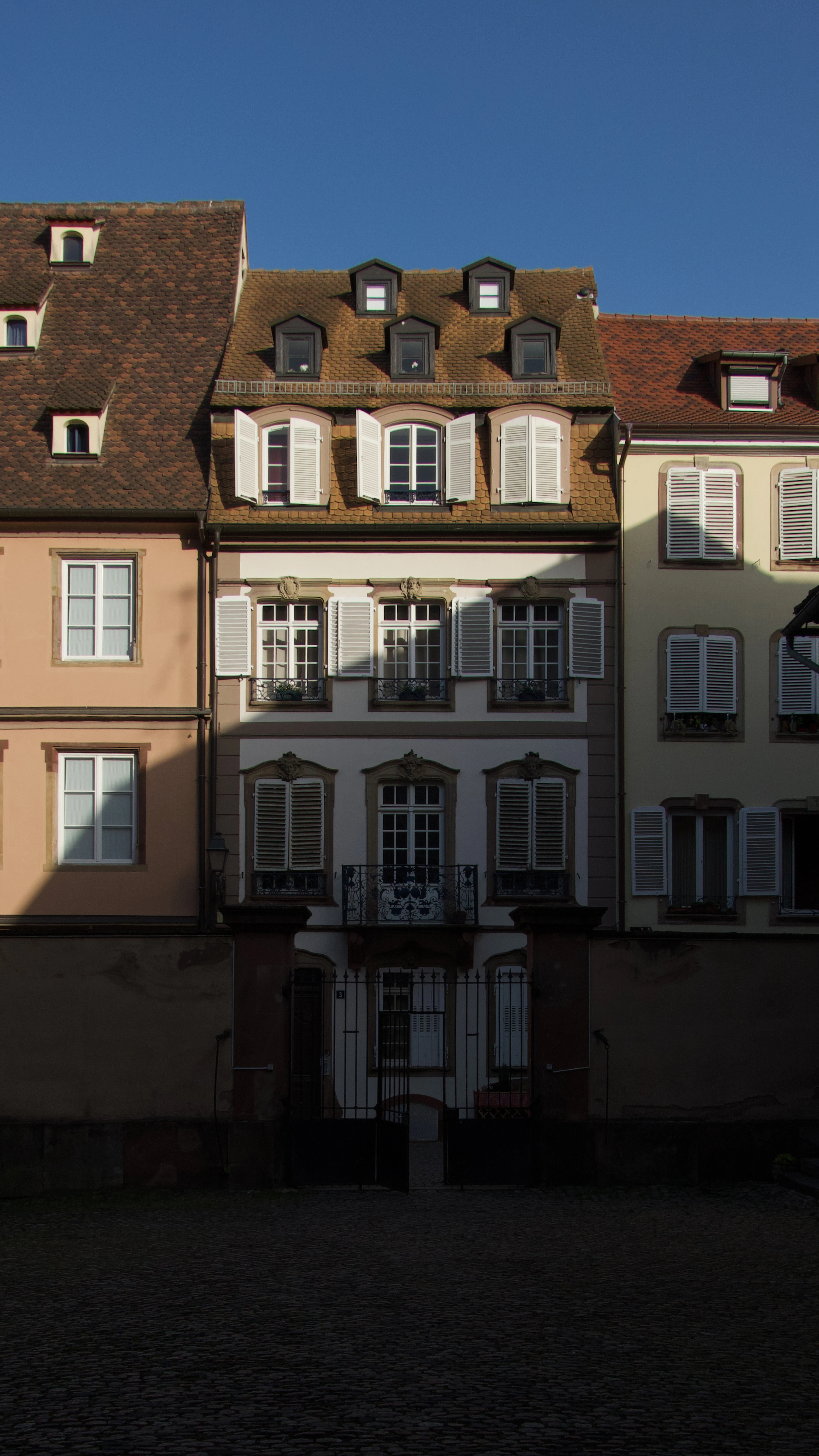Tall eighteenth-century townhouses in Strasbourg with white shutters, dormer windows in steep tiled roofs and a wrought-iron balcony, catching low sun above a cobbled square.