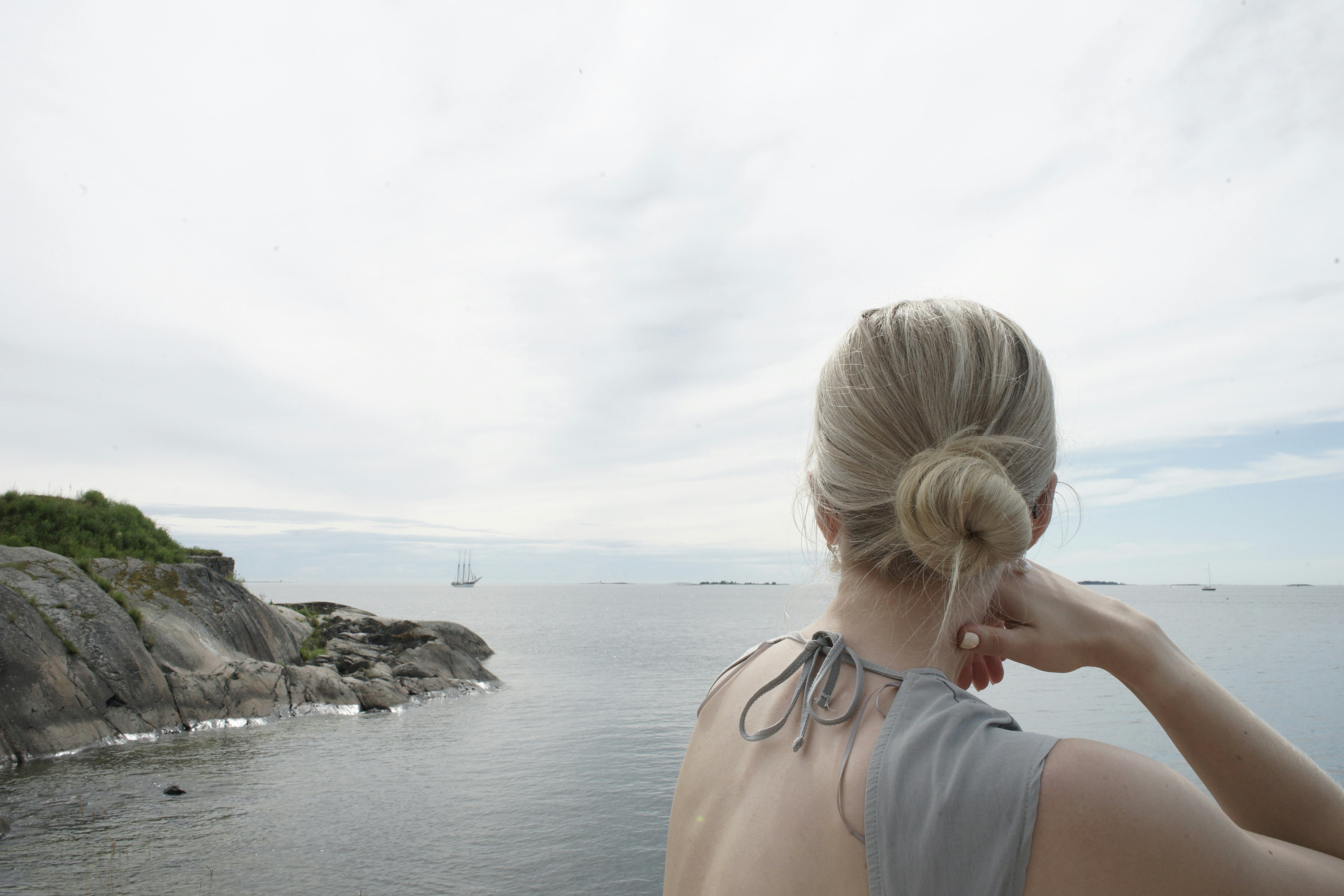 Beautiful blonde woman, with a bare back, staring off into at the open ocean under cloudy skies, spotting an old sailing ship.