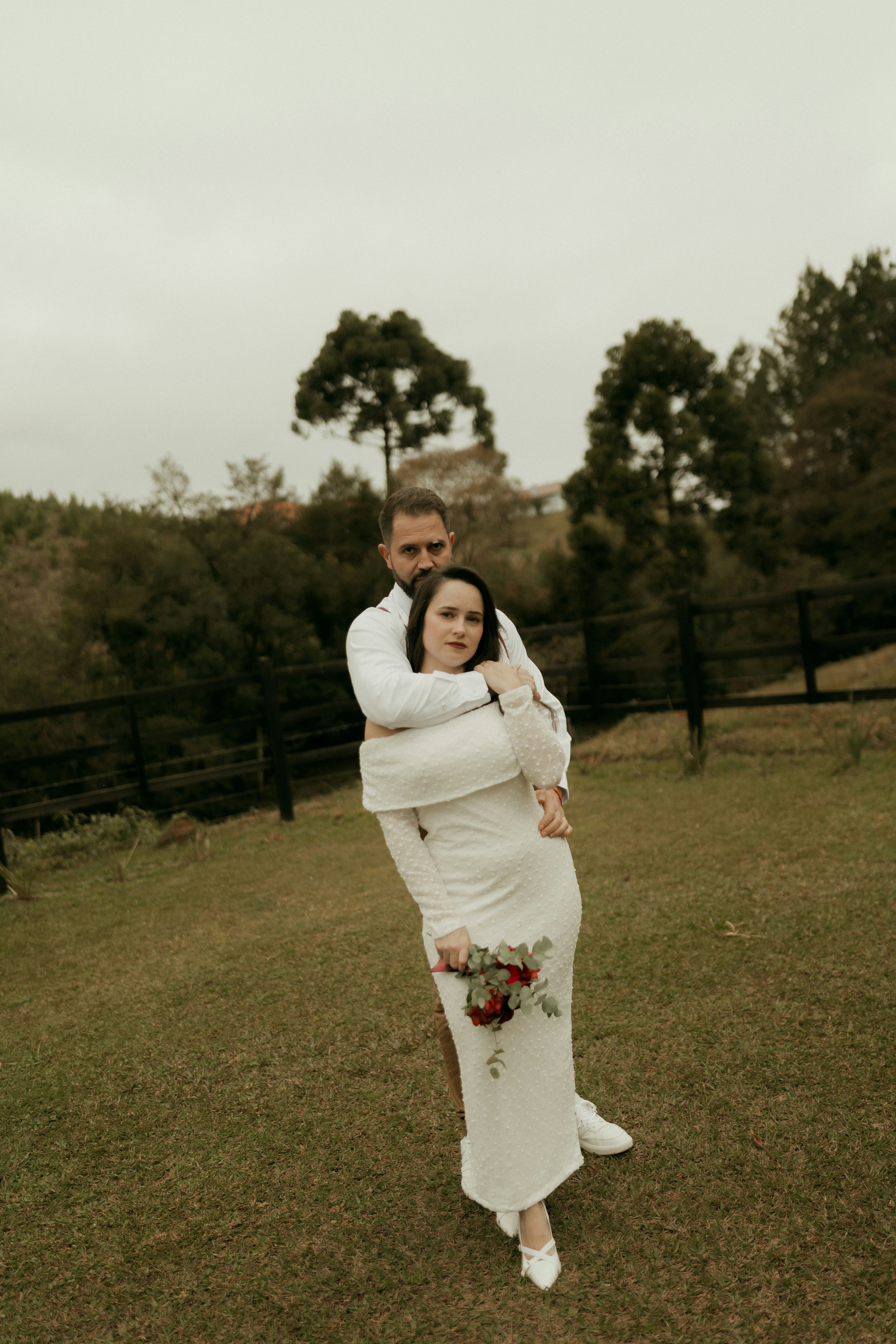 Man embracing woman in white dress with bouquet outdoors.