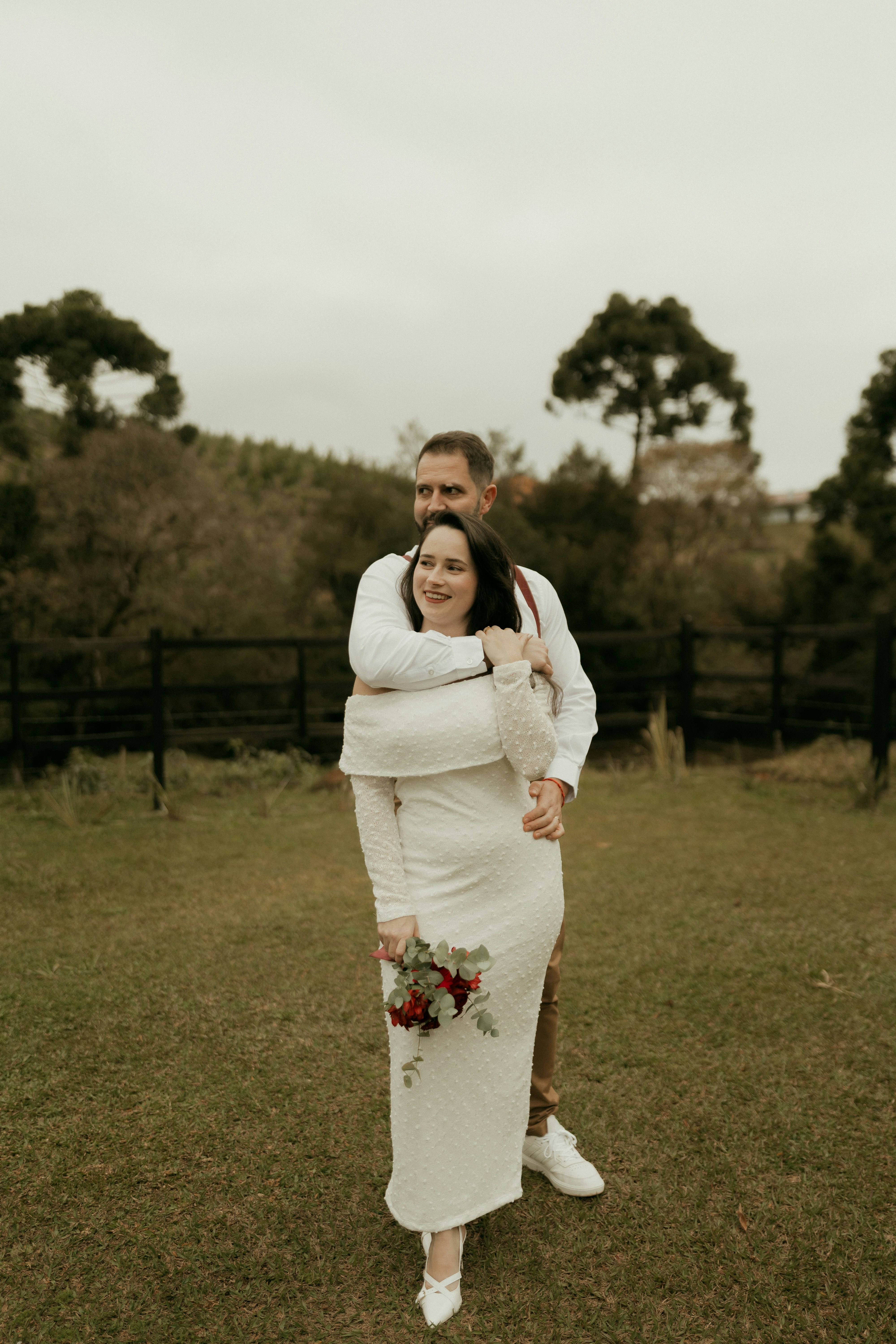 Man embraces woman in white dress holding a bouquet outdoors.