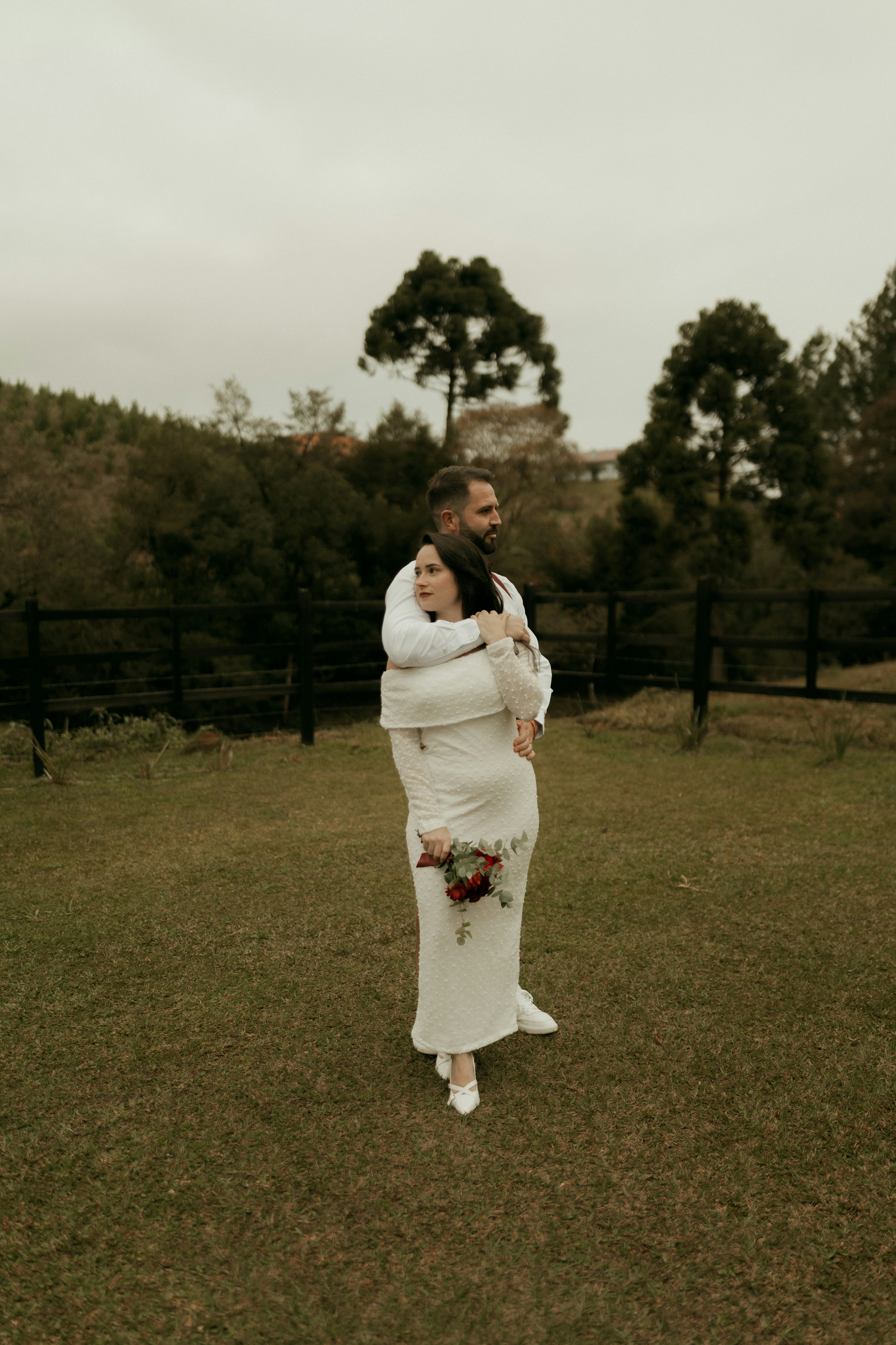 Man hugging woman in white dress holding flowers outdoors.
