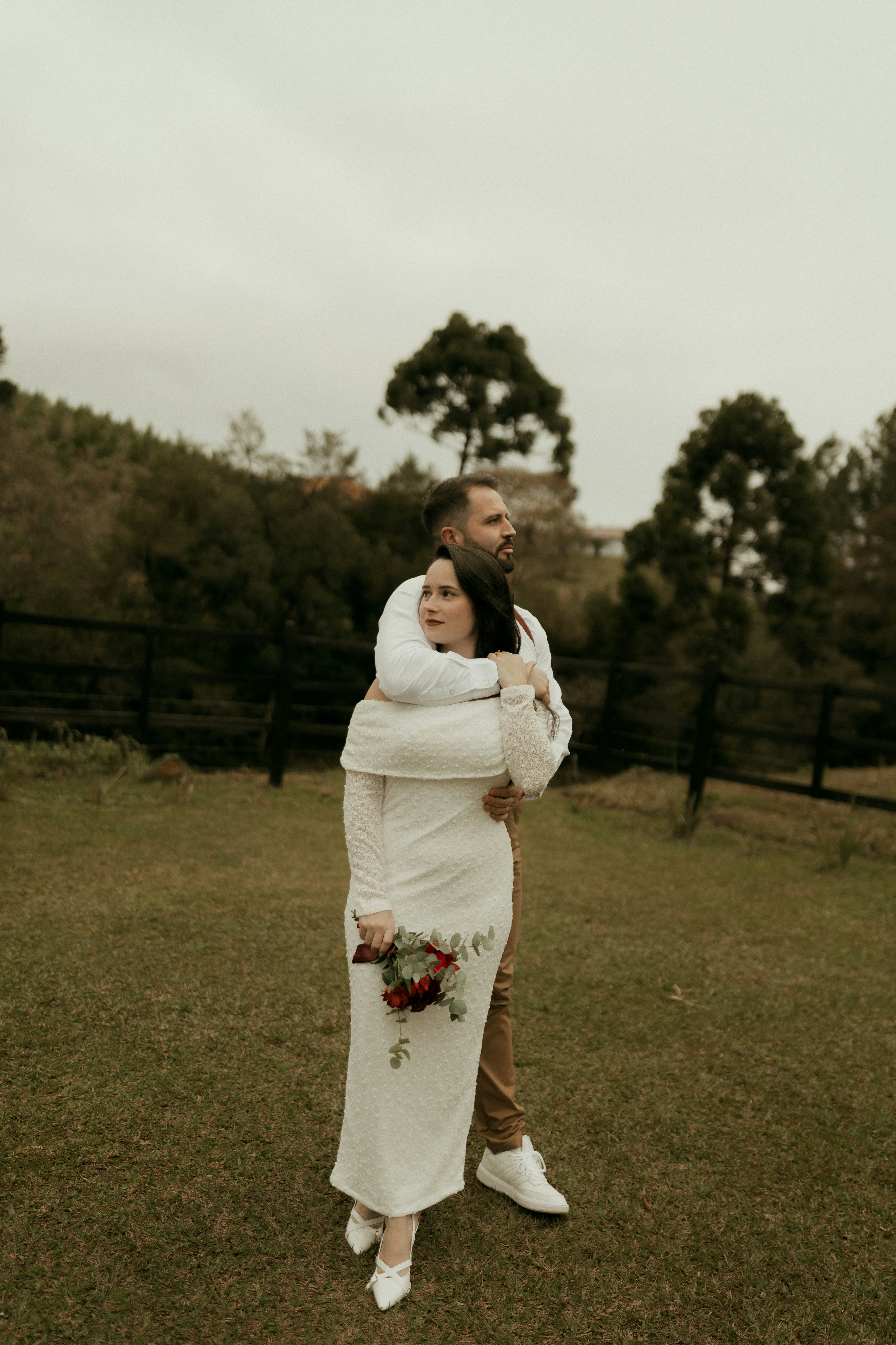 Man embraces woman in white dress holding red flowers outdoors.