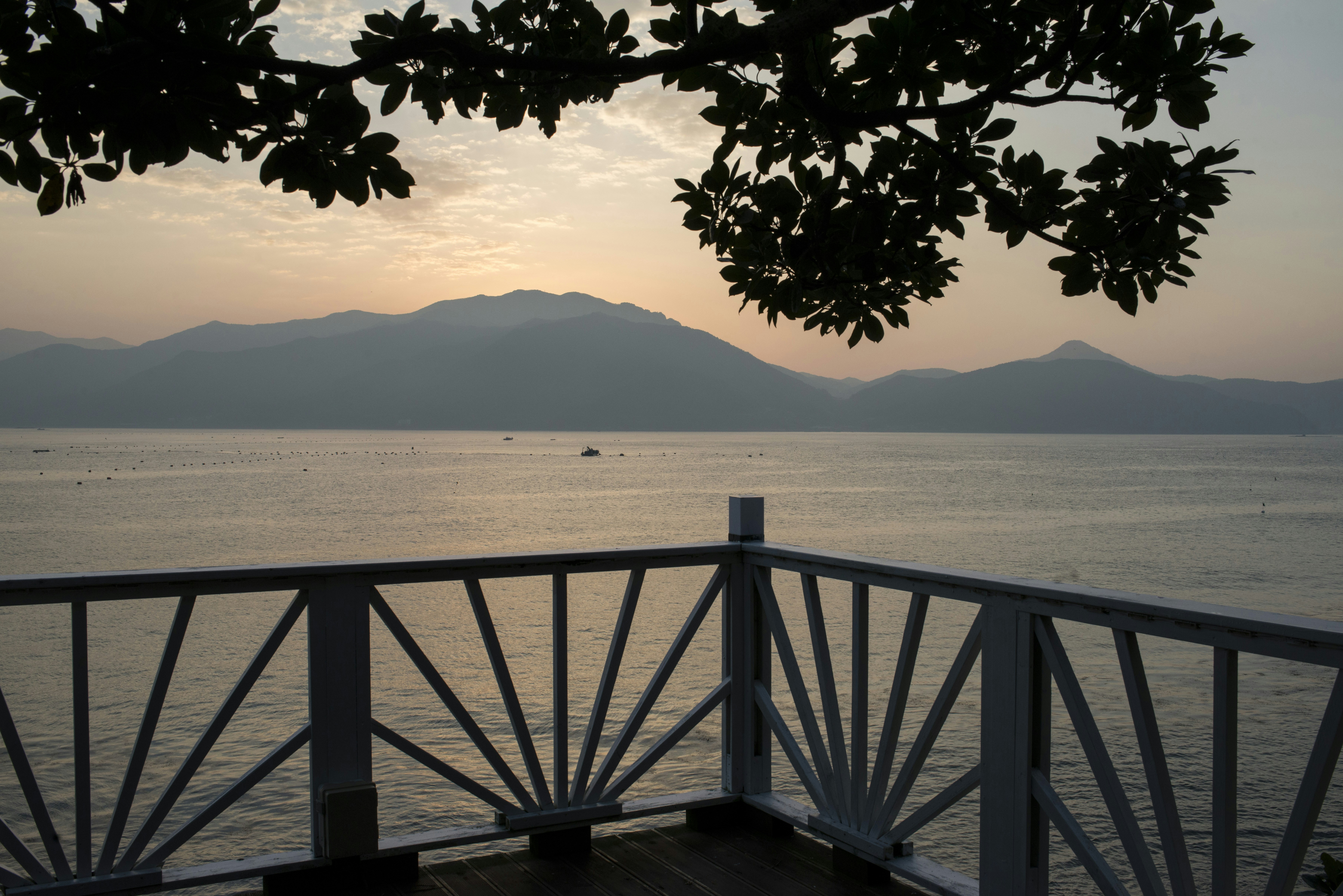 Calm water at sunset with mountains, railing, and silhouetted trees
