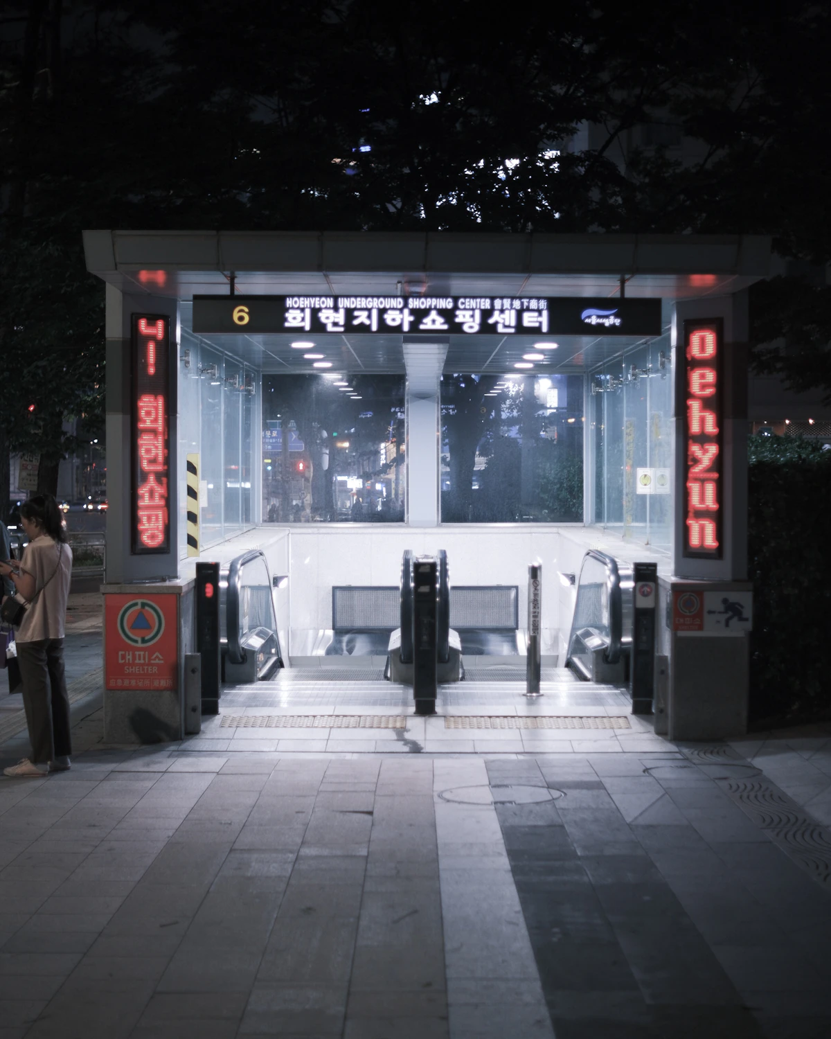 Lit subway station entrance with escalators going down at night, one person standing at the side