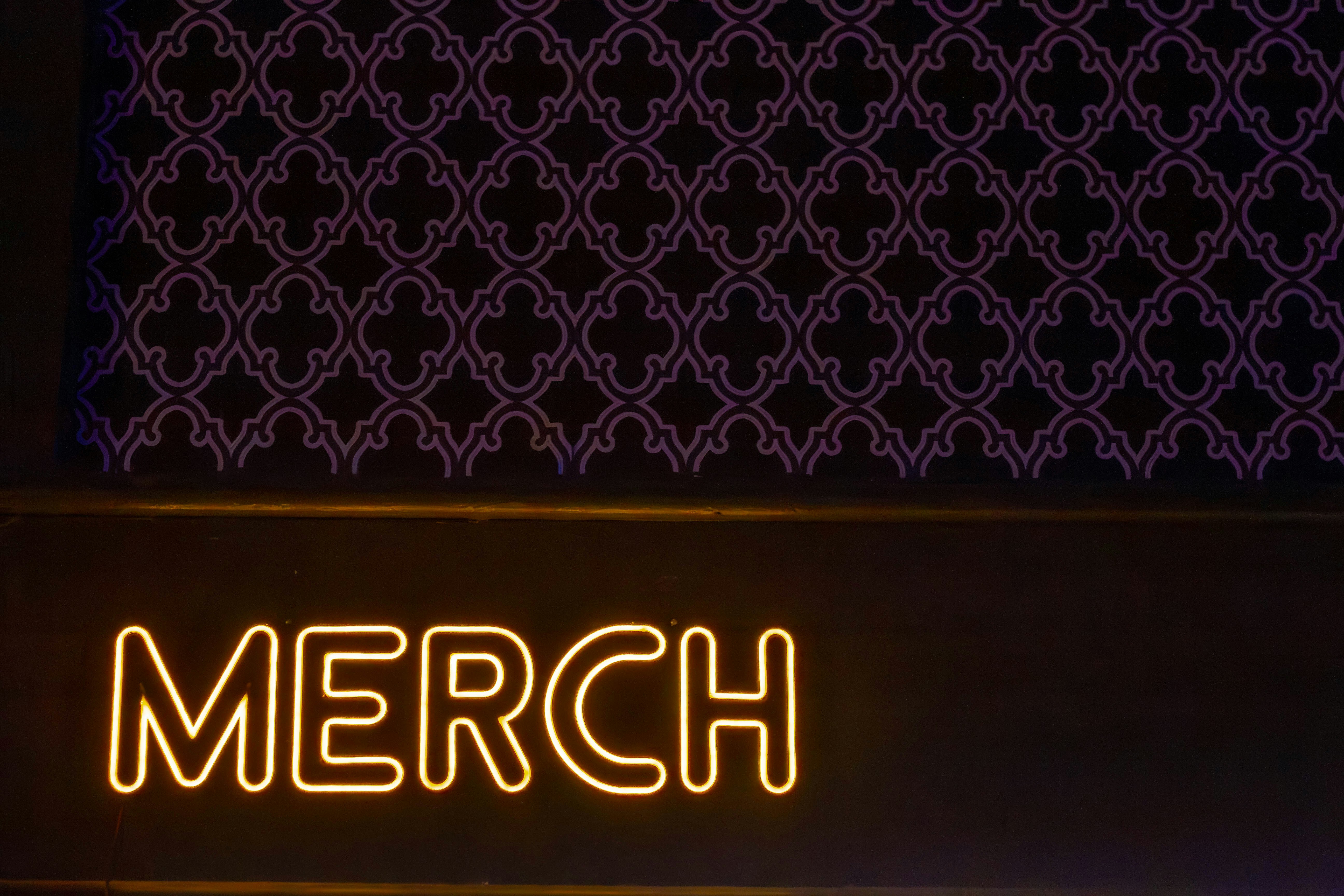 A neon "merch" sign with a dark patterned background.