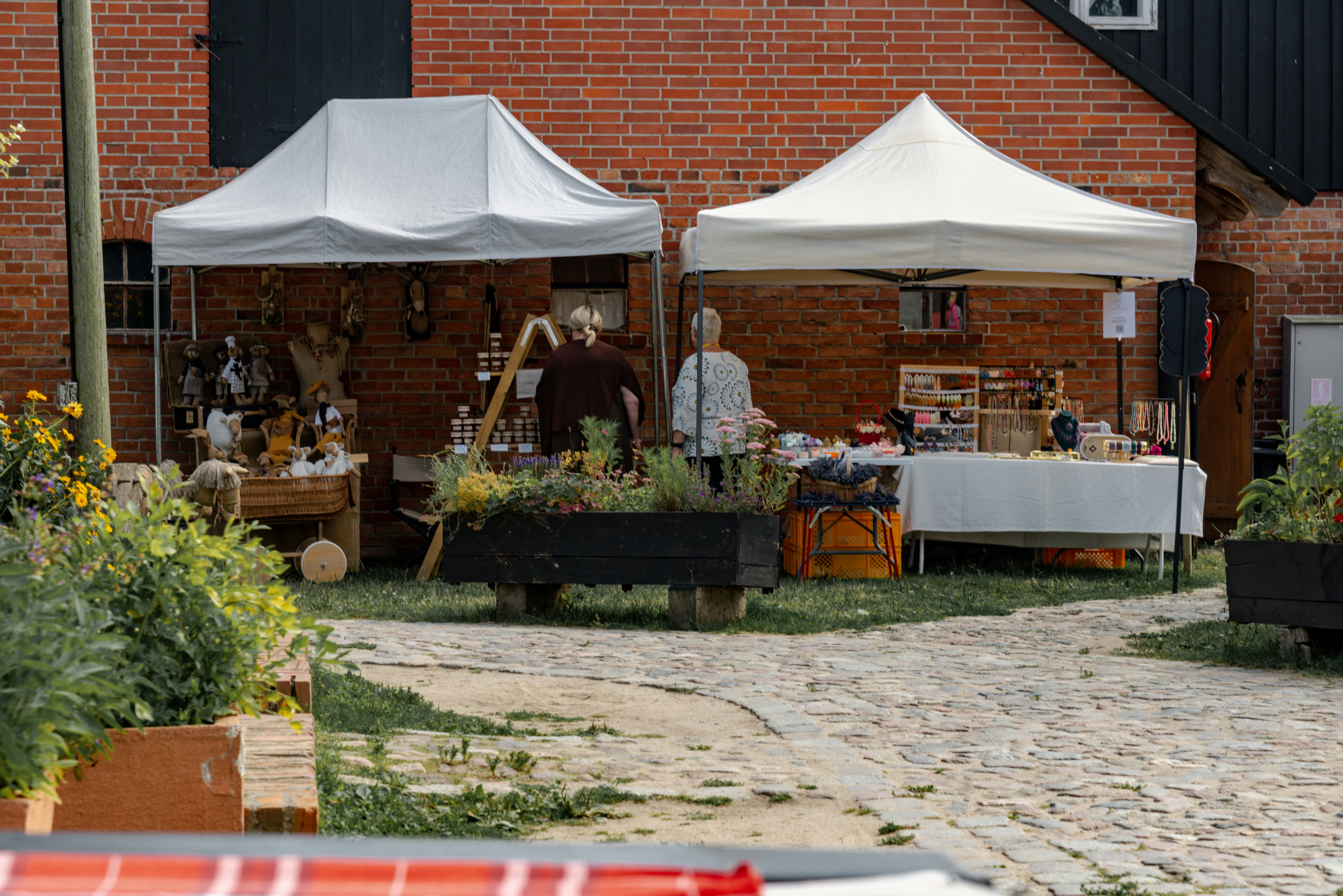 Two market tents display goods outside a rustic brick building.