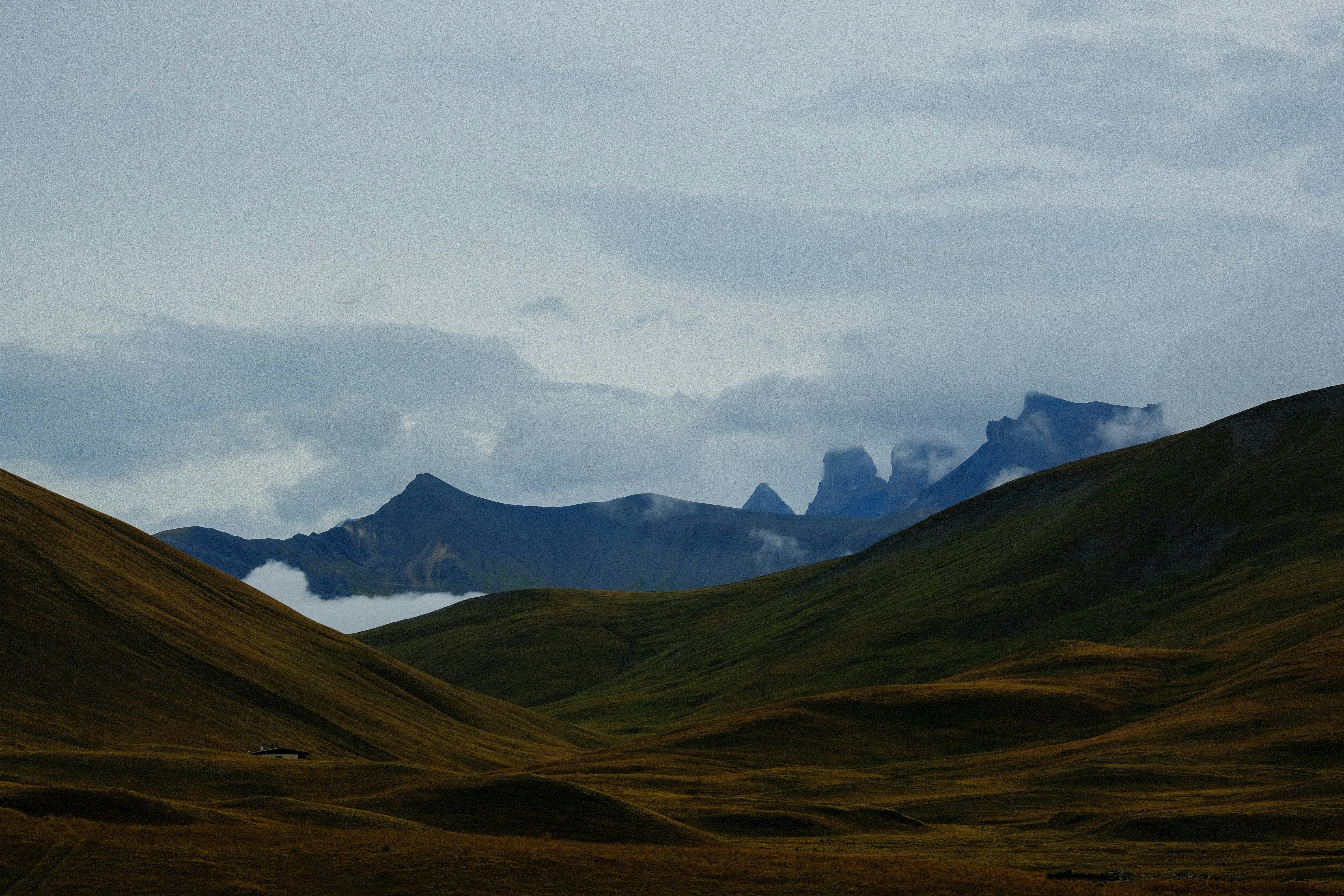 Green hills lead to distant jagged mountains under cloudy sky