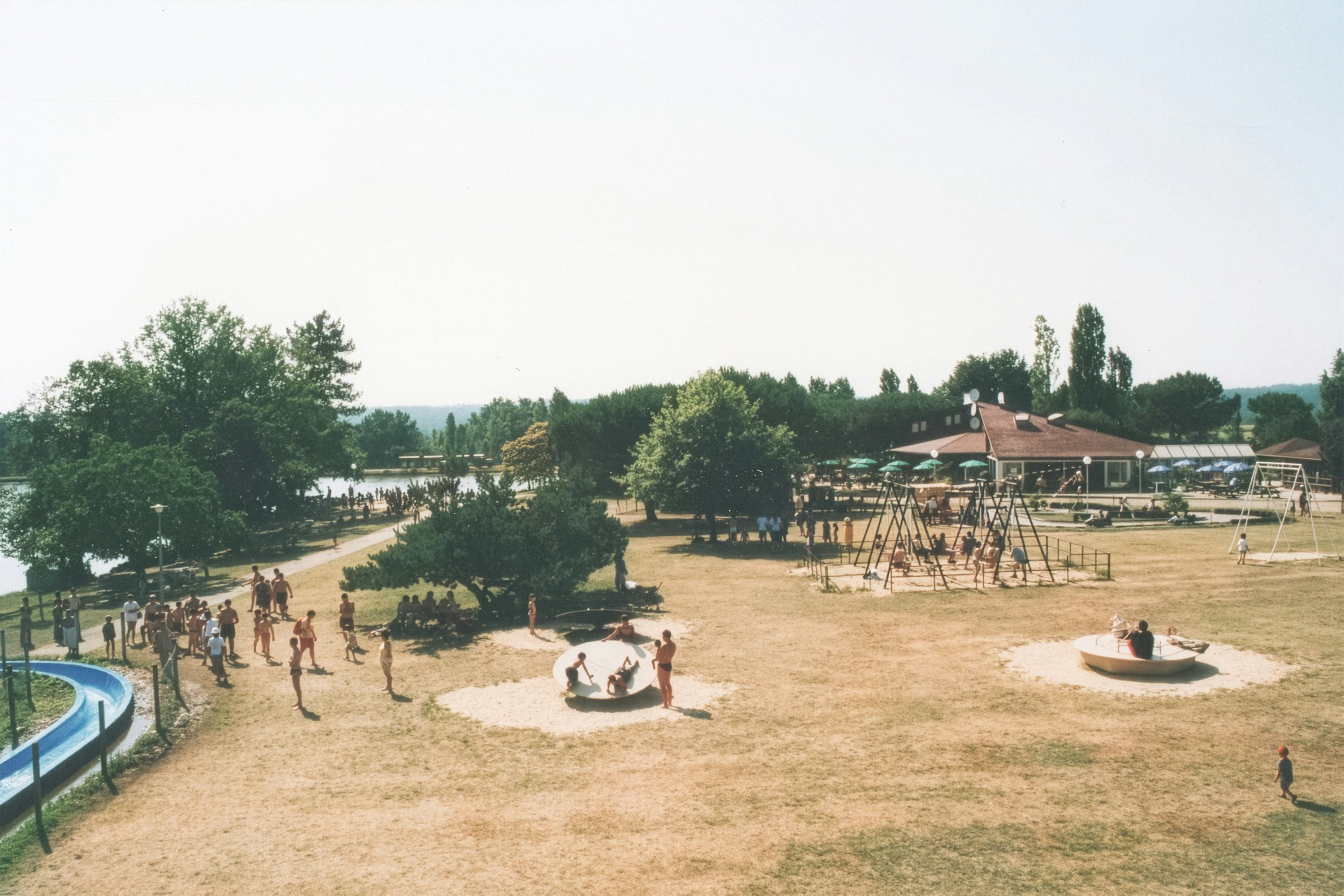 Sunny lakeside park with playground, building, and people.