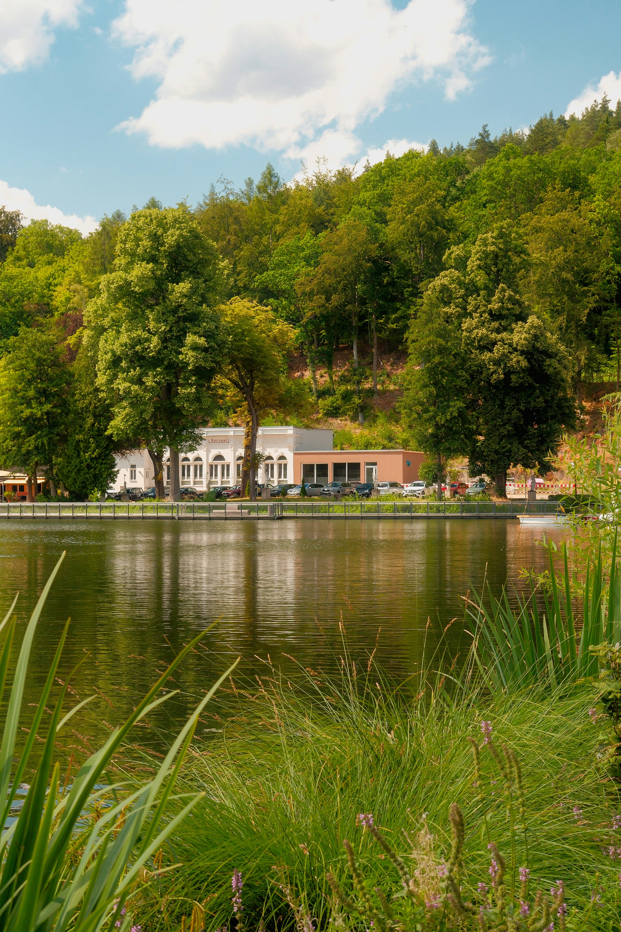 Serene lake reflecting buildings and a lush, tree-covered hillside.