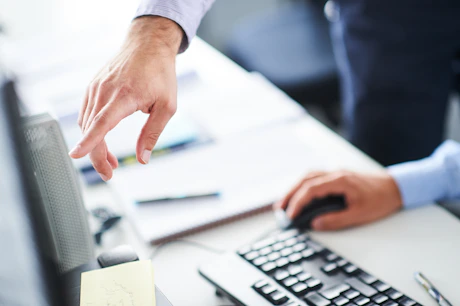 Hands pointing at computer monitor while operating mouse and keyboard.