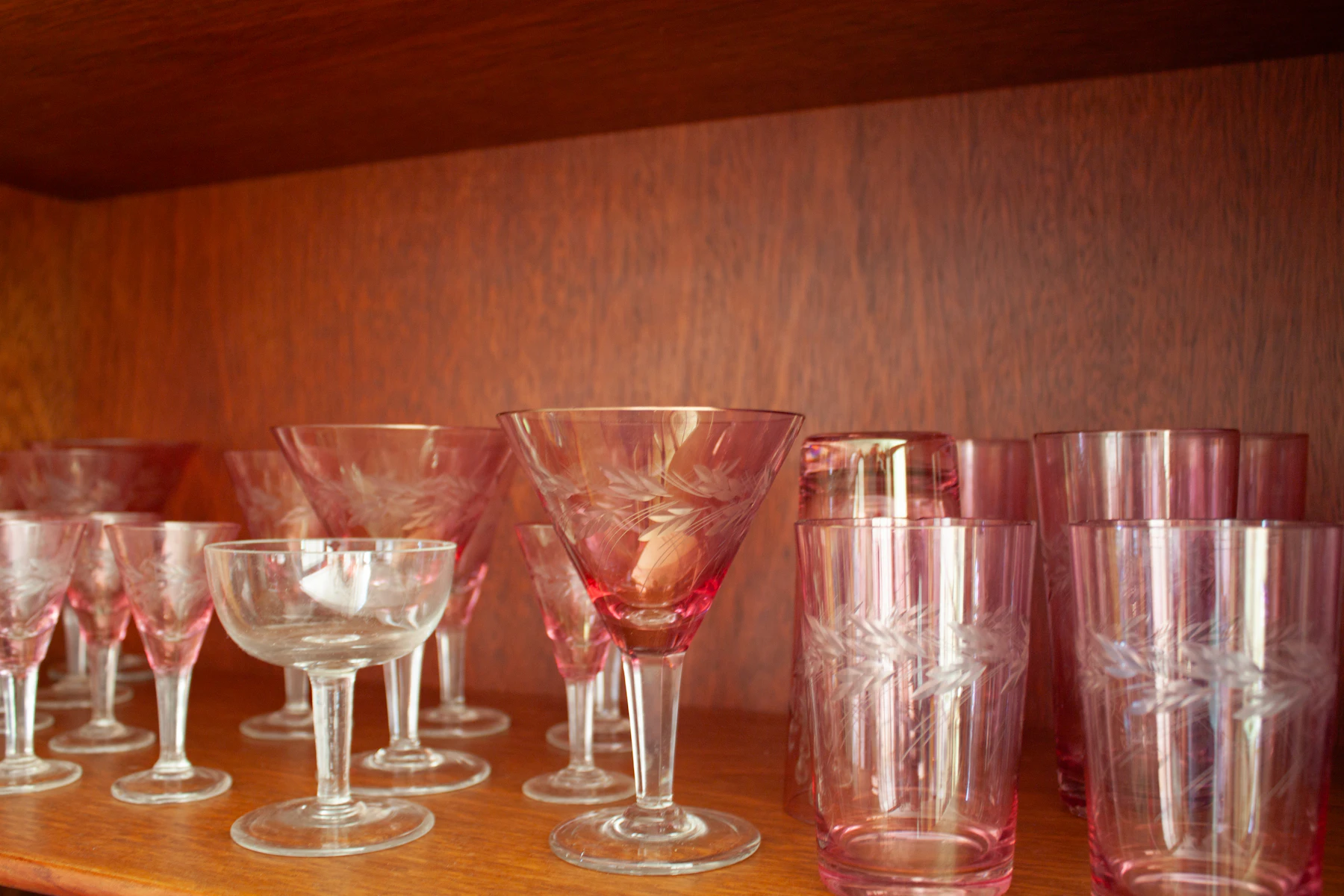 Pink and clear etched stemware and tumblers lined up on a wooden cabinet shelf