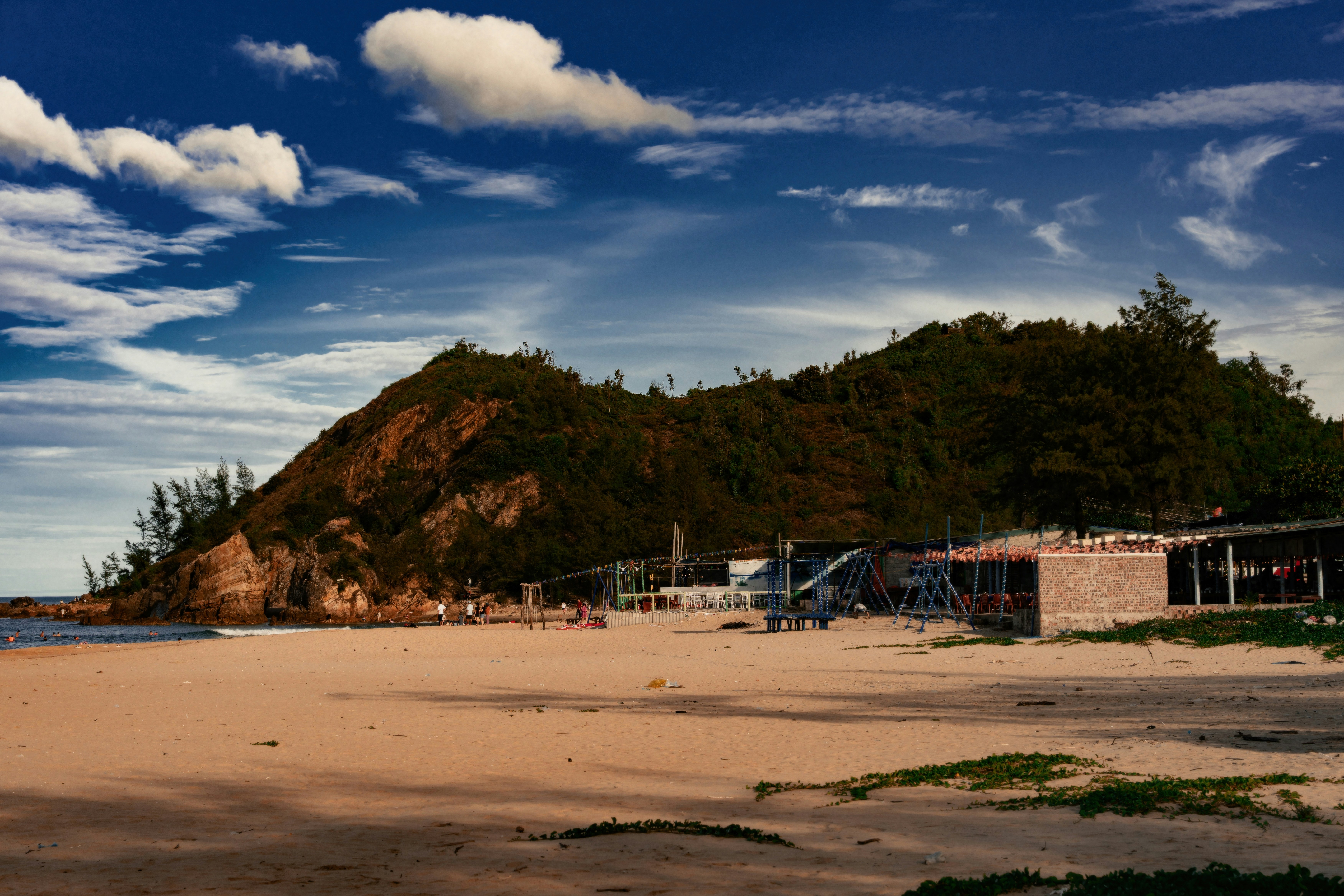 Sandy beach, lush hill, and buildings under a blue sky.