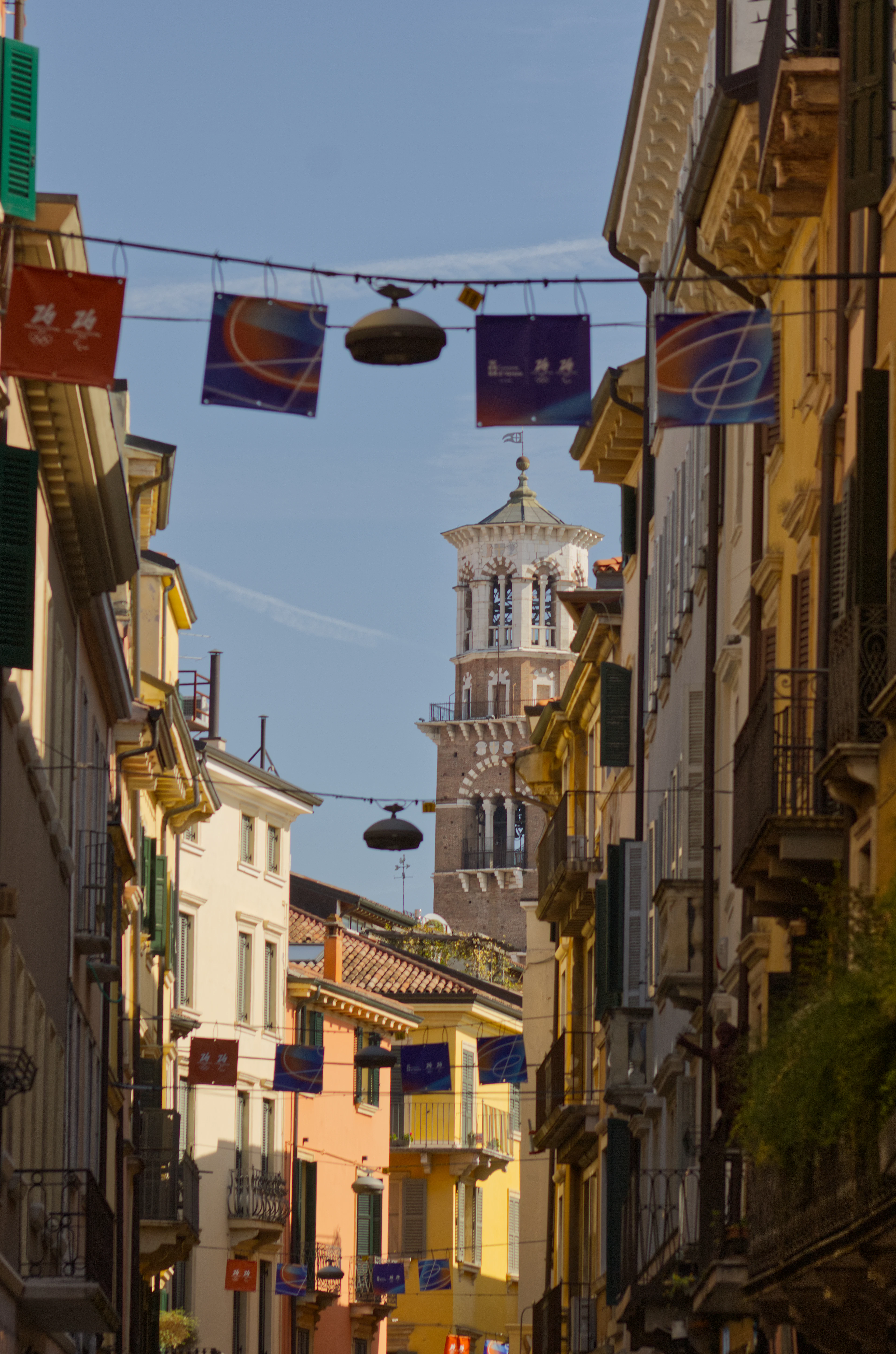 A narrow Verona lane of pastel shuttered houses and iron balconies strung with banners, the white marble belfry of the Torre dei Lamberti closing the view at the far end under a bright blue sky.