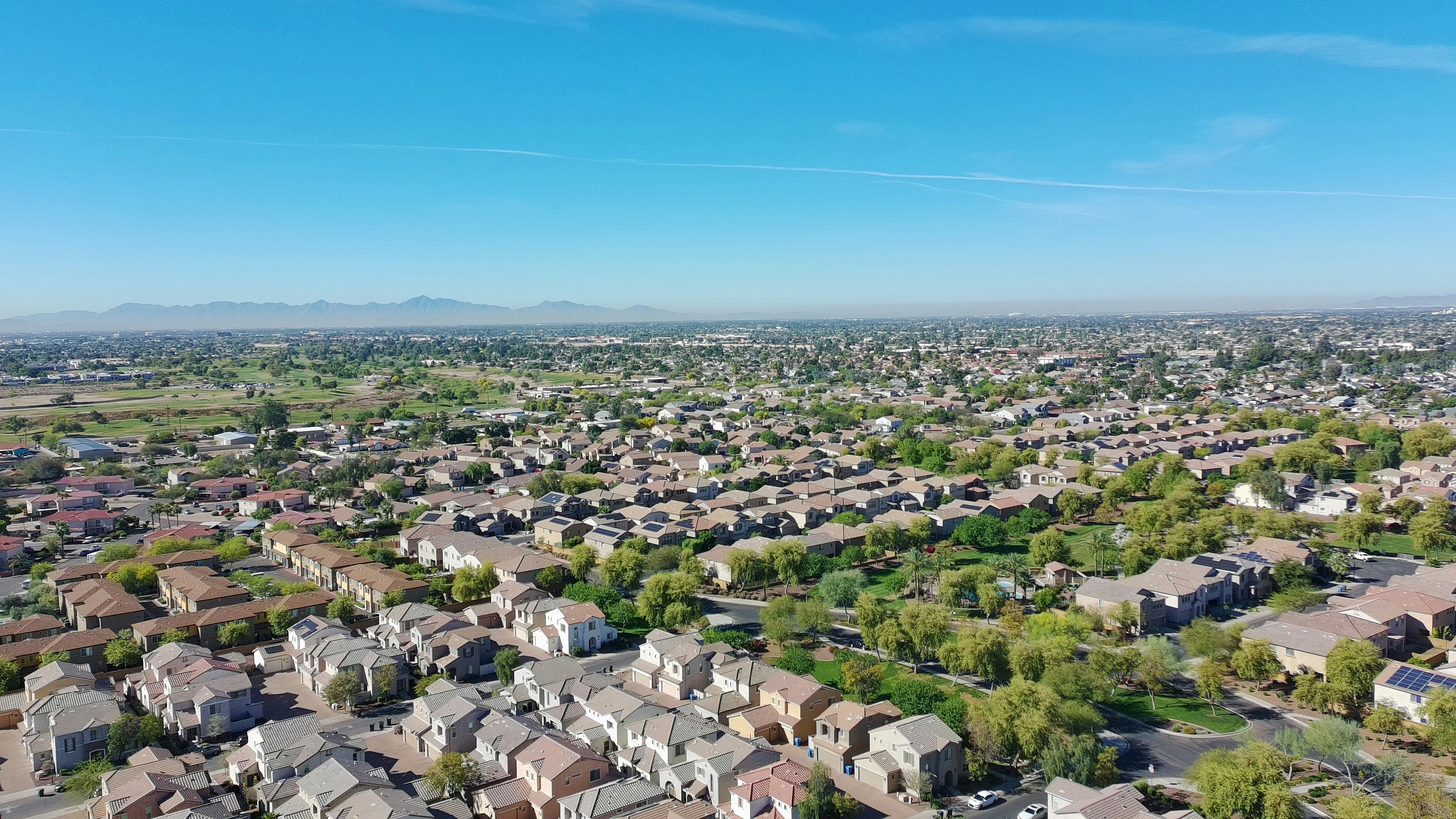 A wide view of a sunlit suburb with rows of houses, streets, and patches of greenery stretching toward distant hills.