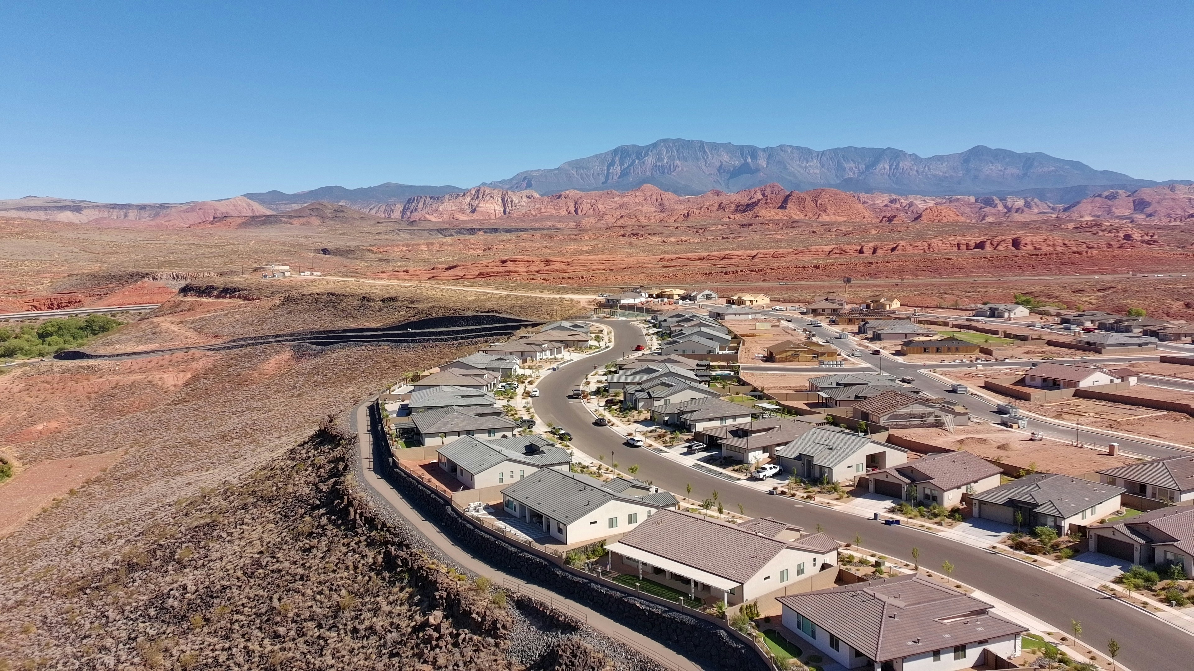 A winding cul-de-sac of modern homes sits at the edge of a desert plain, with red rock formations rising on the horizon under clear daylight.