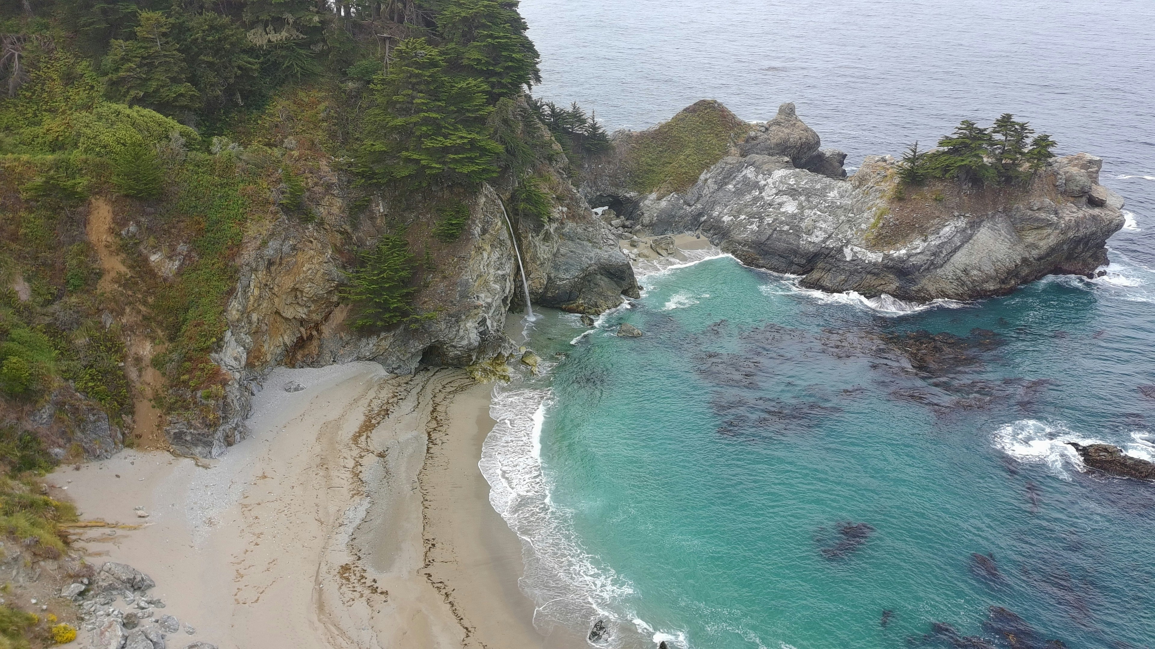 Coastal cove with turquoise water washing onto a sandy beach framed by rocky cliffs and green trees. Natural shoreline scene showcasing contrast between ocean, rock and vegetation.