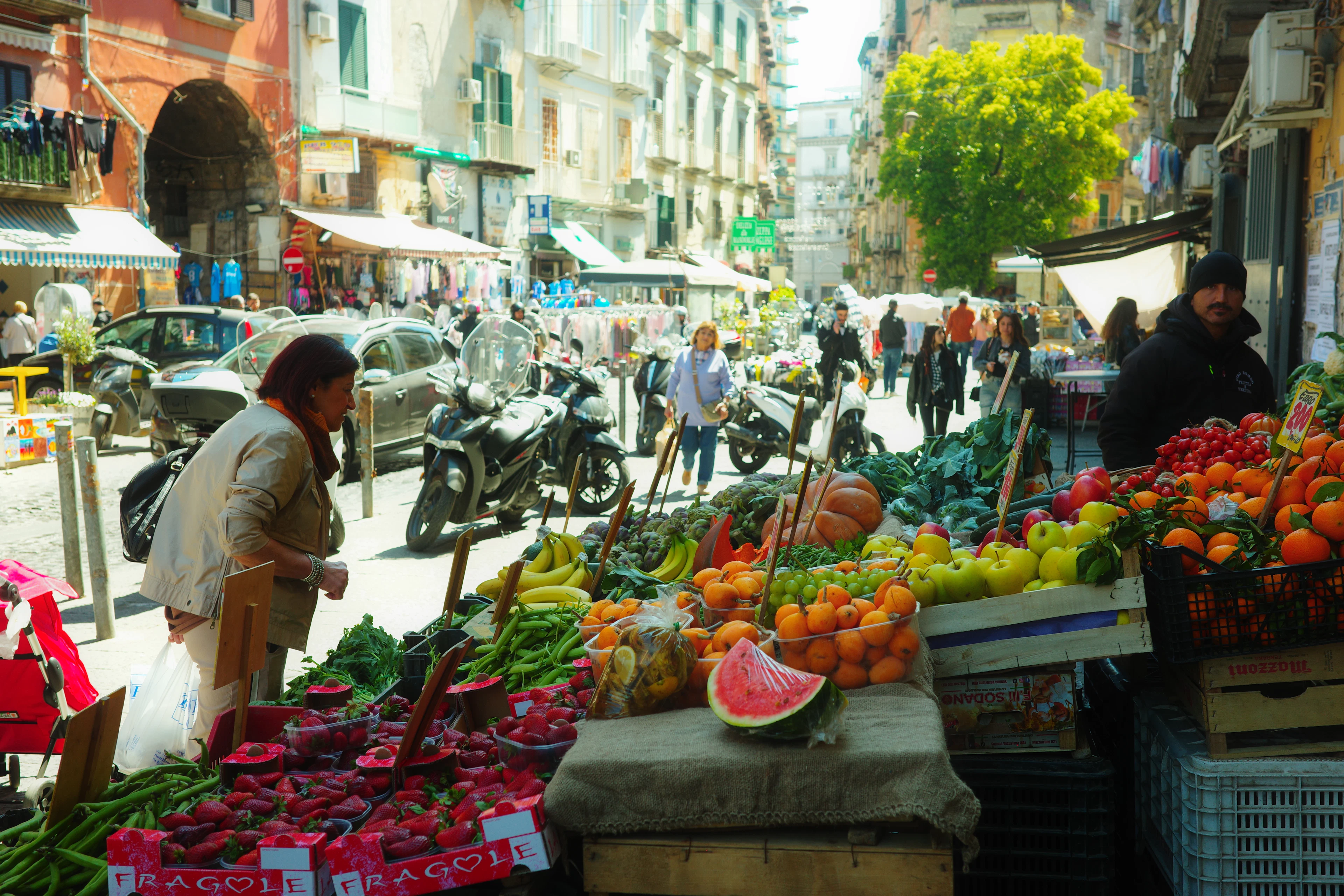 A Naples street market in full swing, trays of strawberries, oranges, peppers and greens spilling onto the pavement beside a cut watermelon while shoppers, scooters and parked cars fill the lane behind.