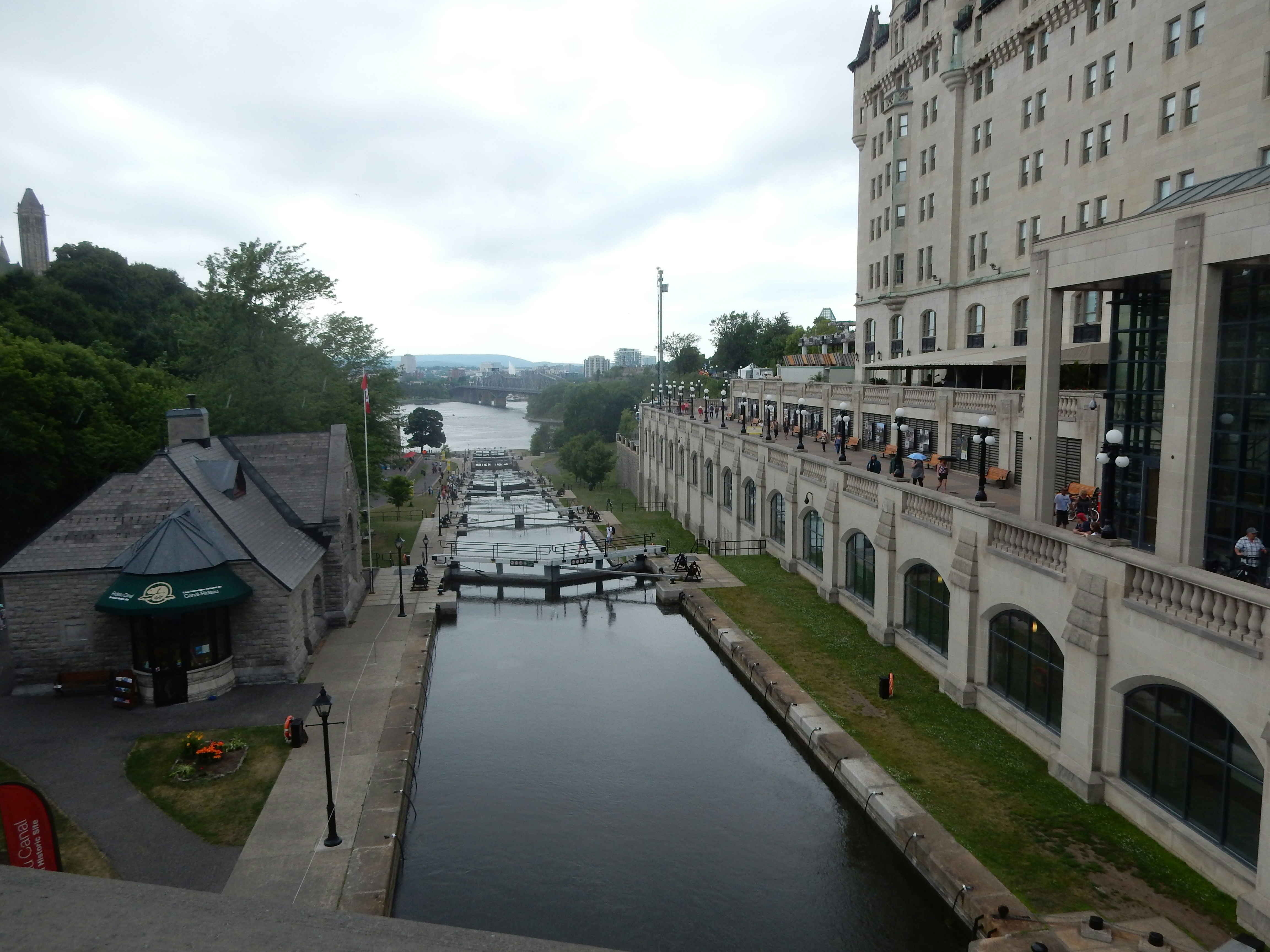 Wide canal flanked by a grand stone building and a riverside promenade, with pedestrians along the terrace and a calm, reflective waterway disappearing into the distance.