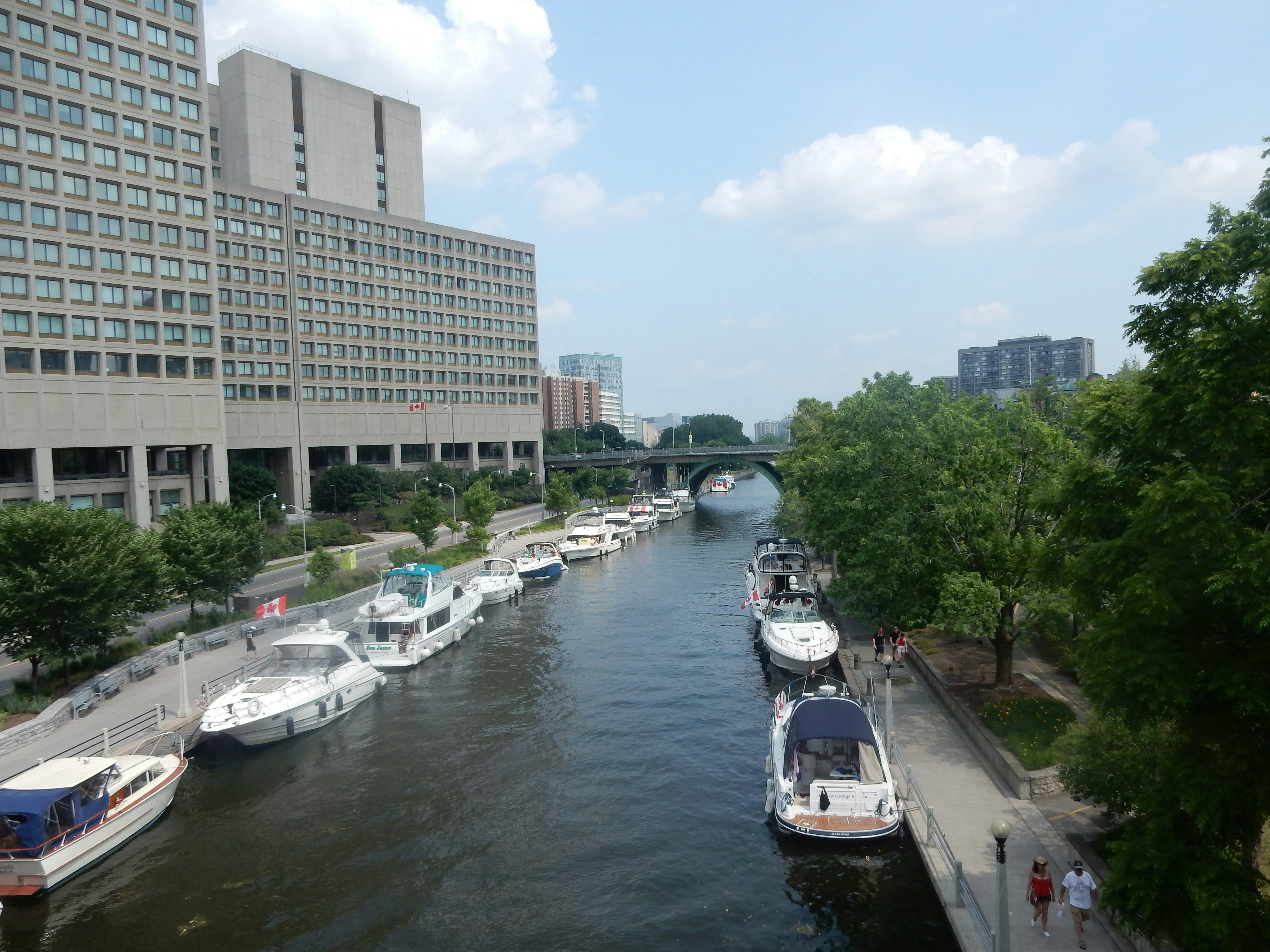 City canal with boats docked along both sides under a blue sky and modern buildings, a bridge in the distance; a serene urban waterway scene.