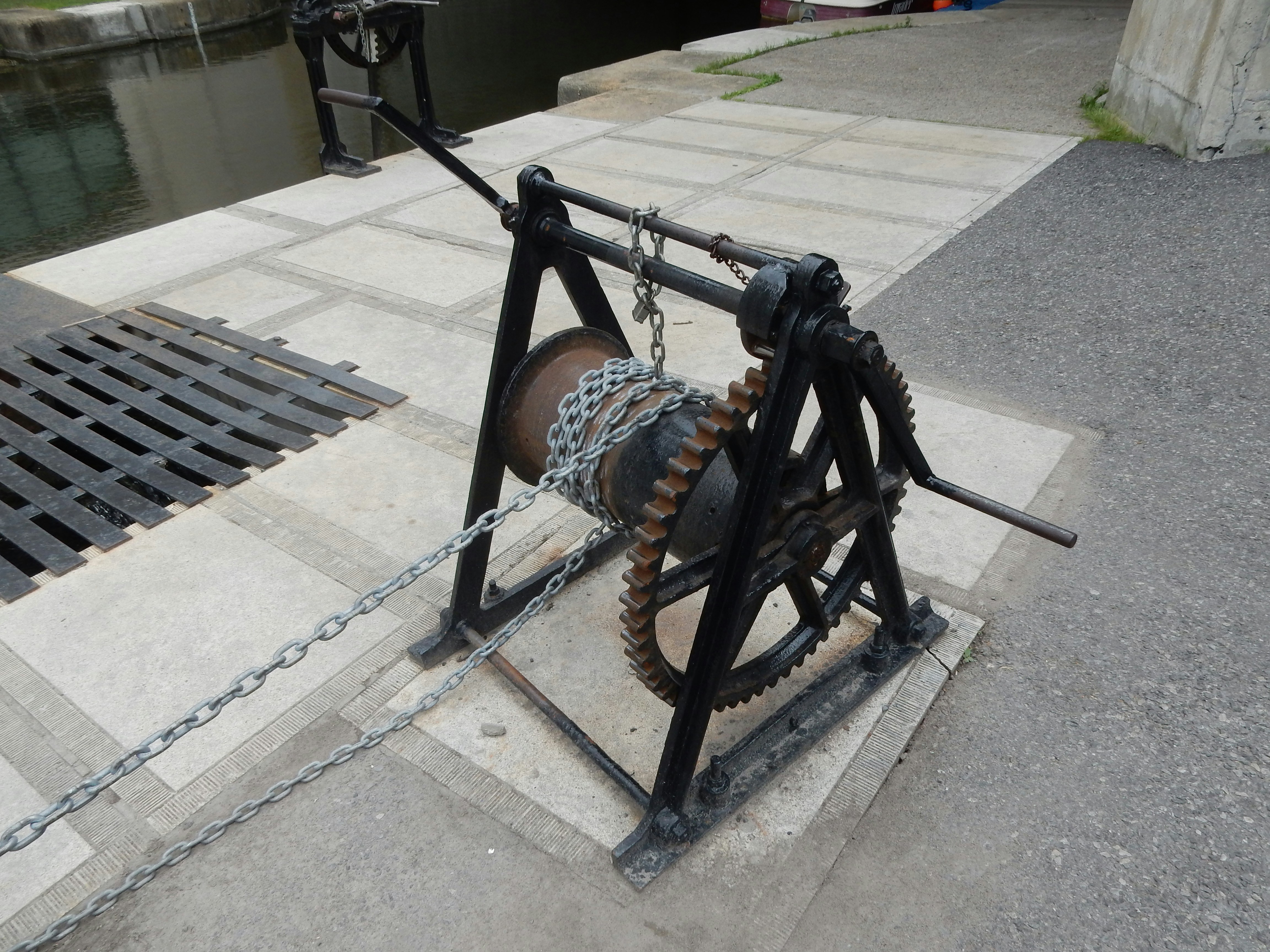 Rusty gear and chain mechanism anchored on a concrete platform beside a canal, captured at street level with surrounding pavement and a grate in view. This is a structural, industrial subject.