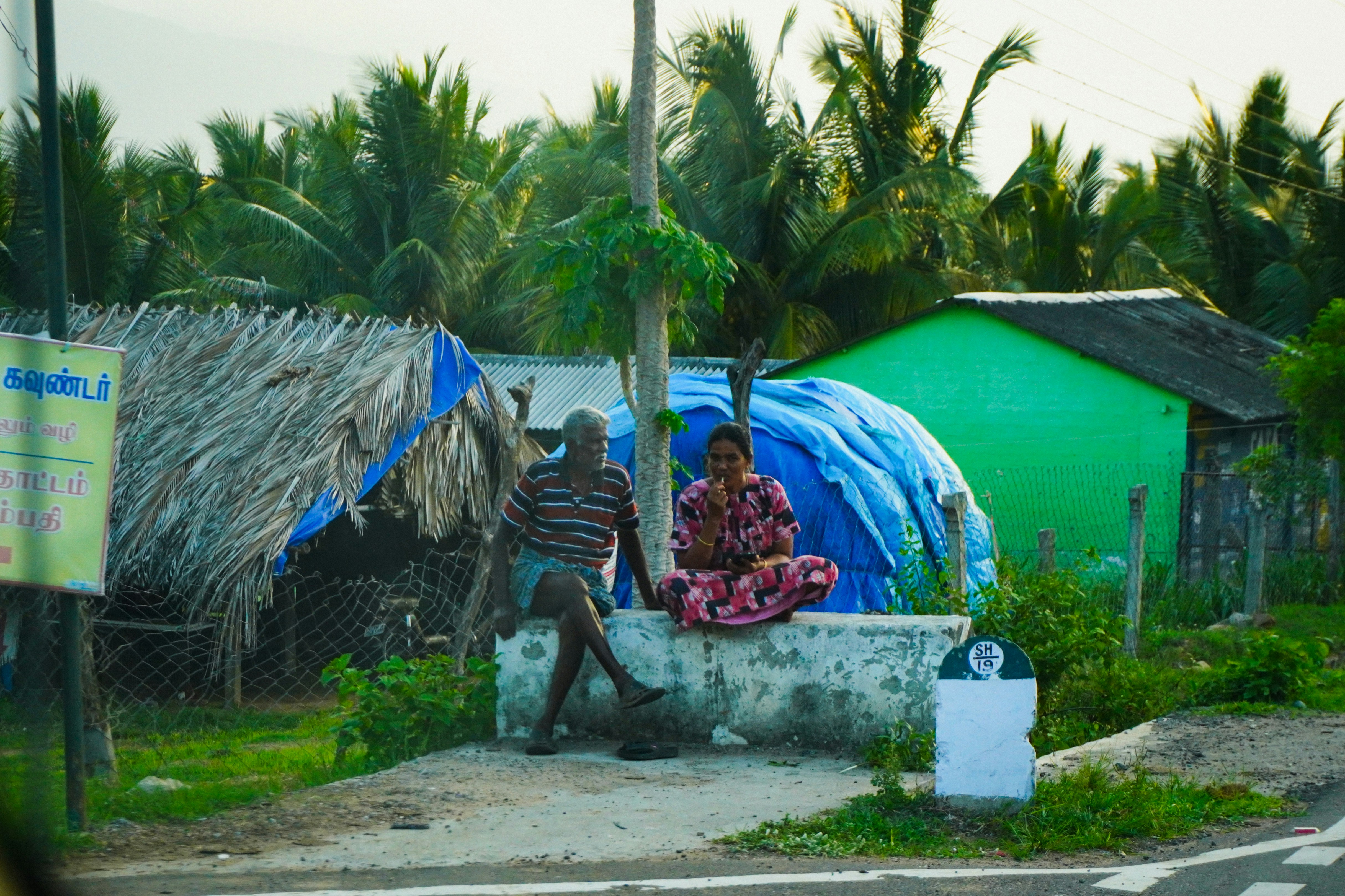 Man and woman sit by roadside near rural huts