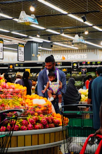 Man carrying baby past vibrant fruit displays in a supermarket