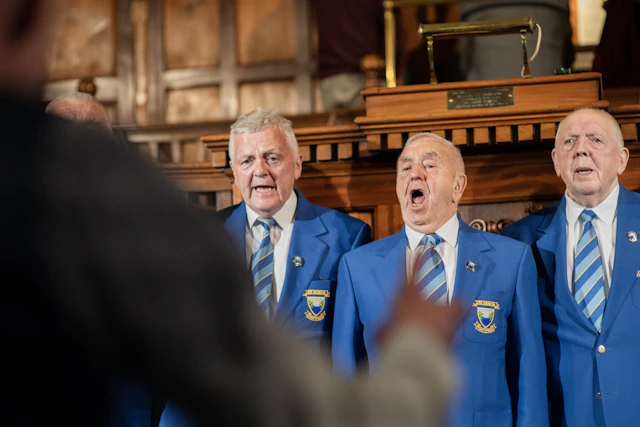 Three older men in blazers singing together indoors
