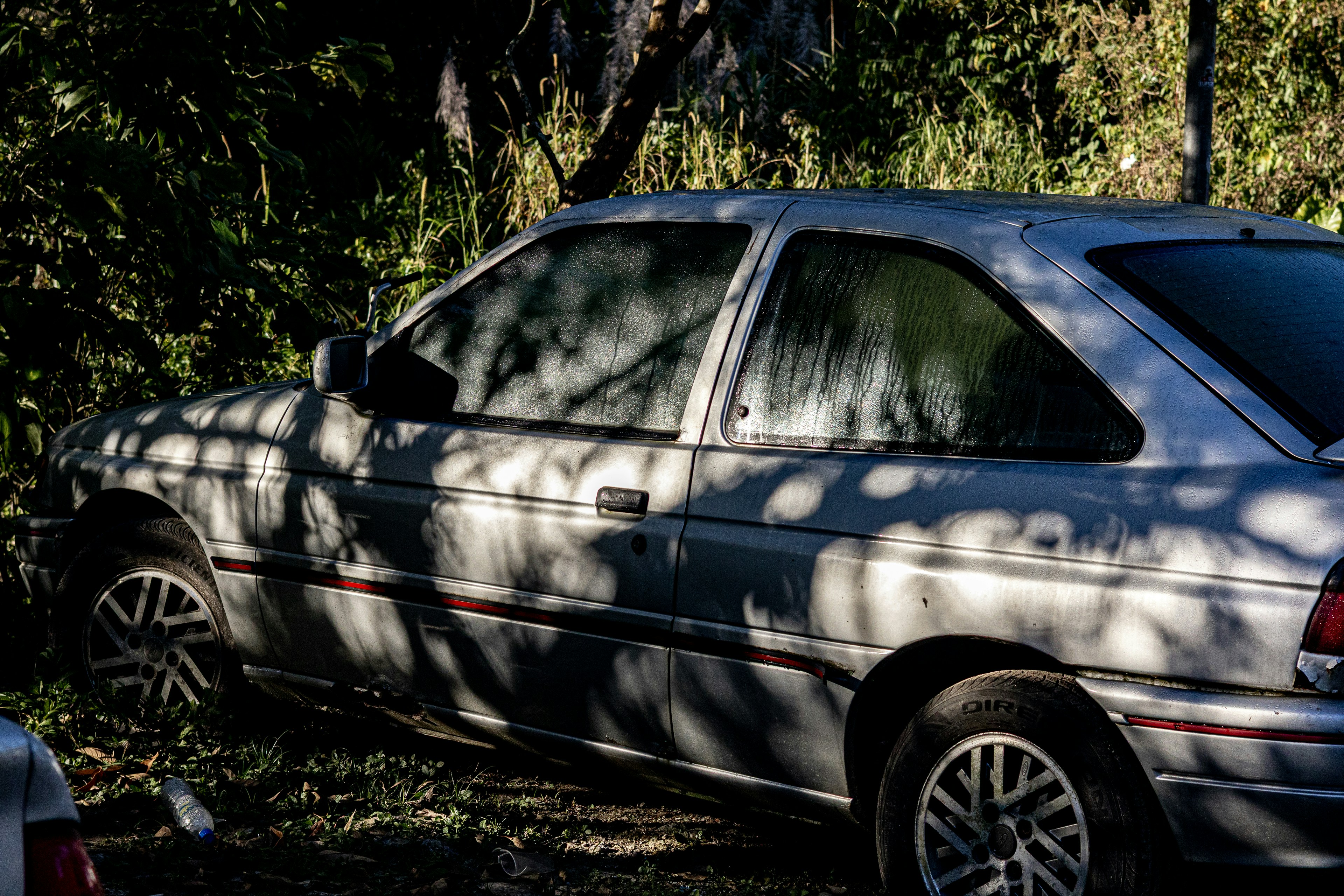 An old silver car parked under dappled tree shadows.