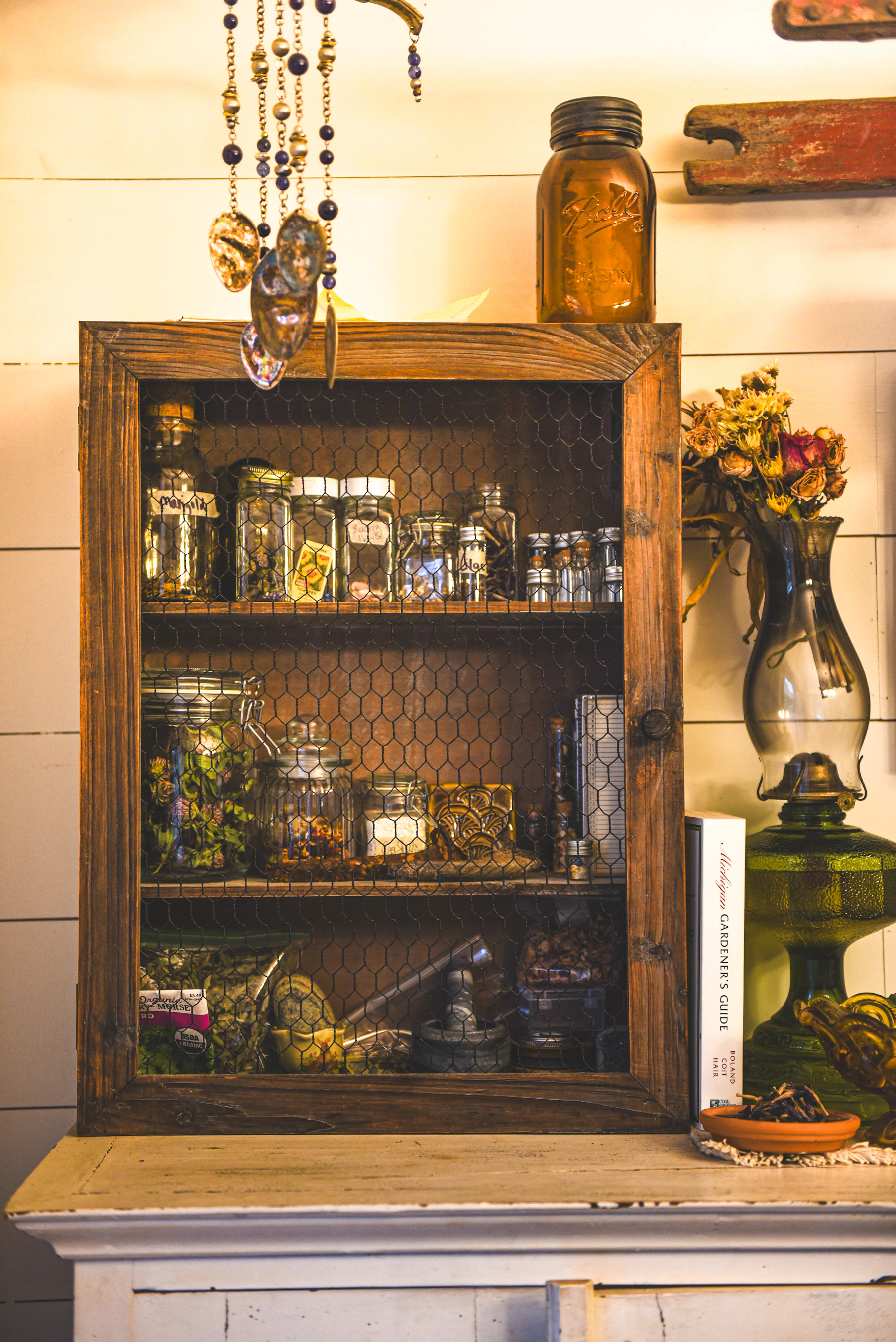 A wooden wall cabinet with a wire door, its shelves full of glass jars of dried herbs