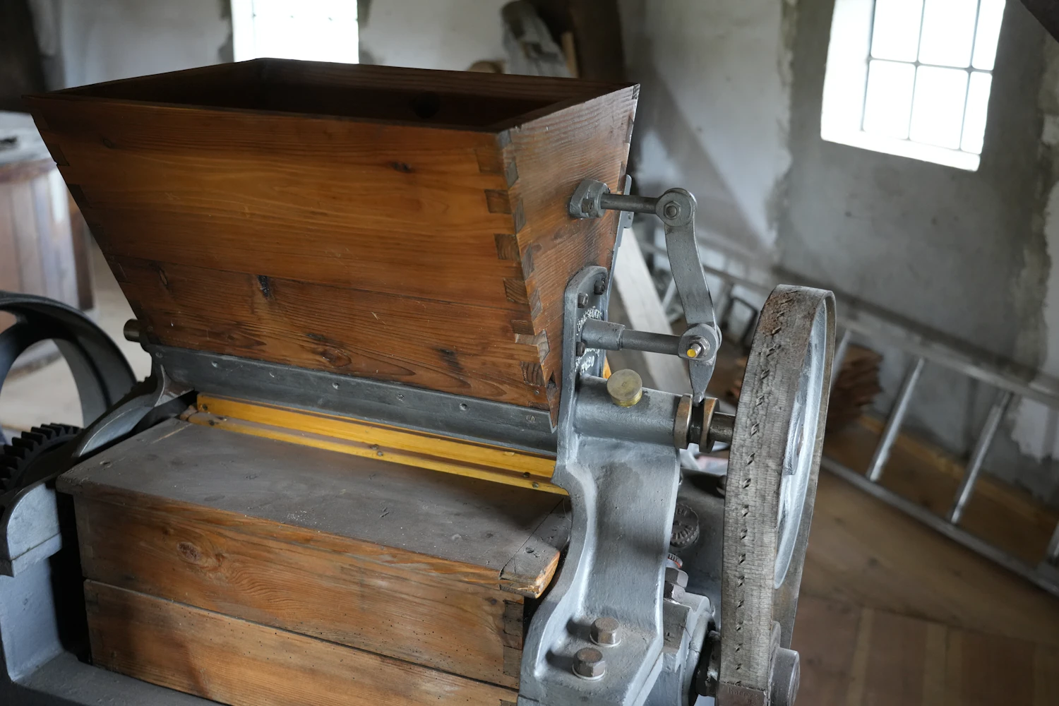 A wooden feed hopper above a cast-iron grain crusher beside a small window