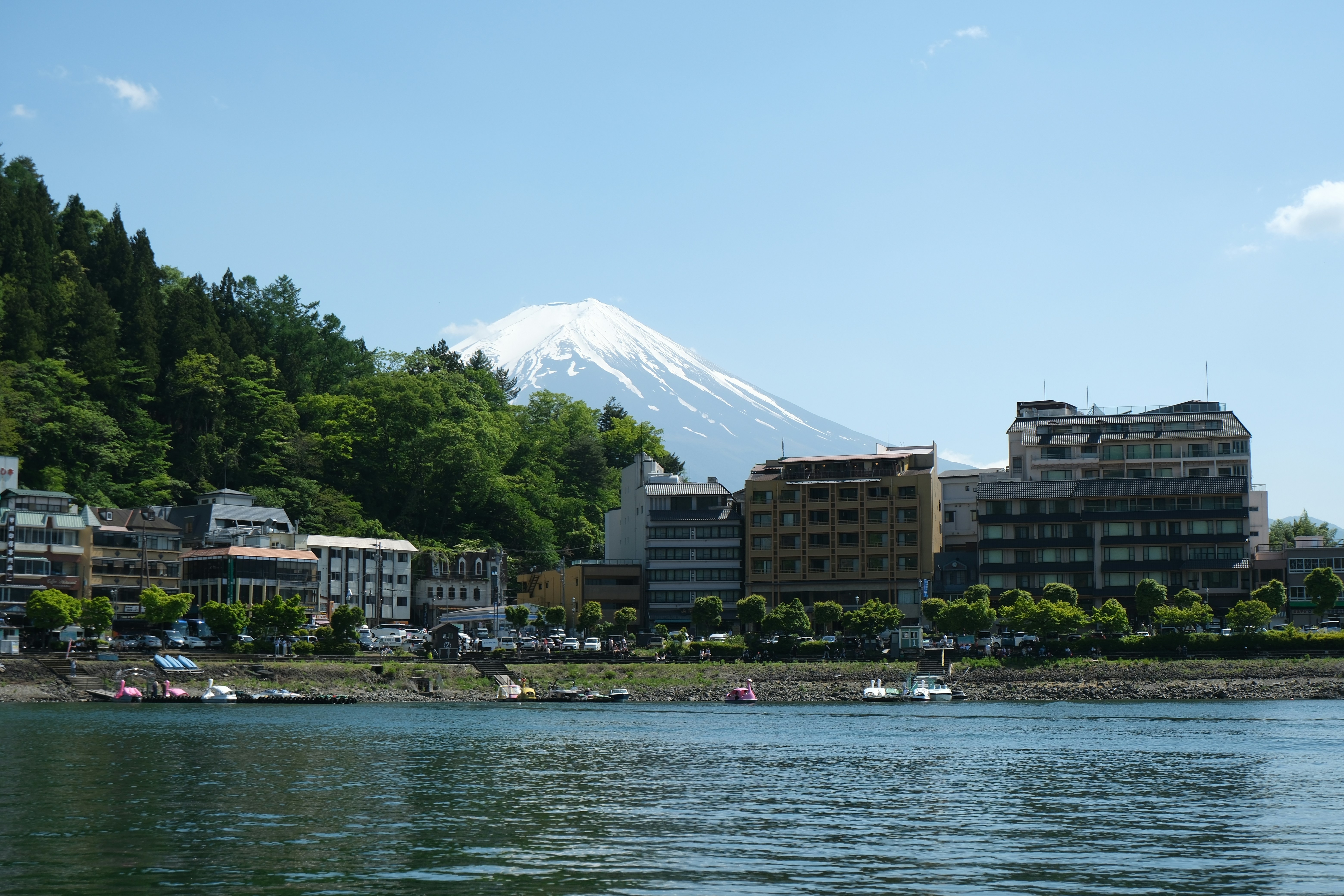Le mont Fuji surplombe une ville au bord d’un lac avec des bâtiments et des bateaux.