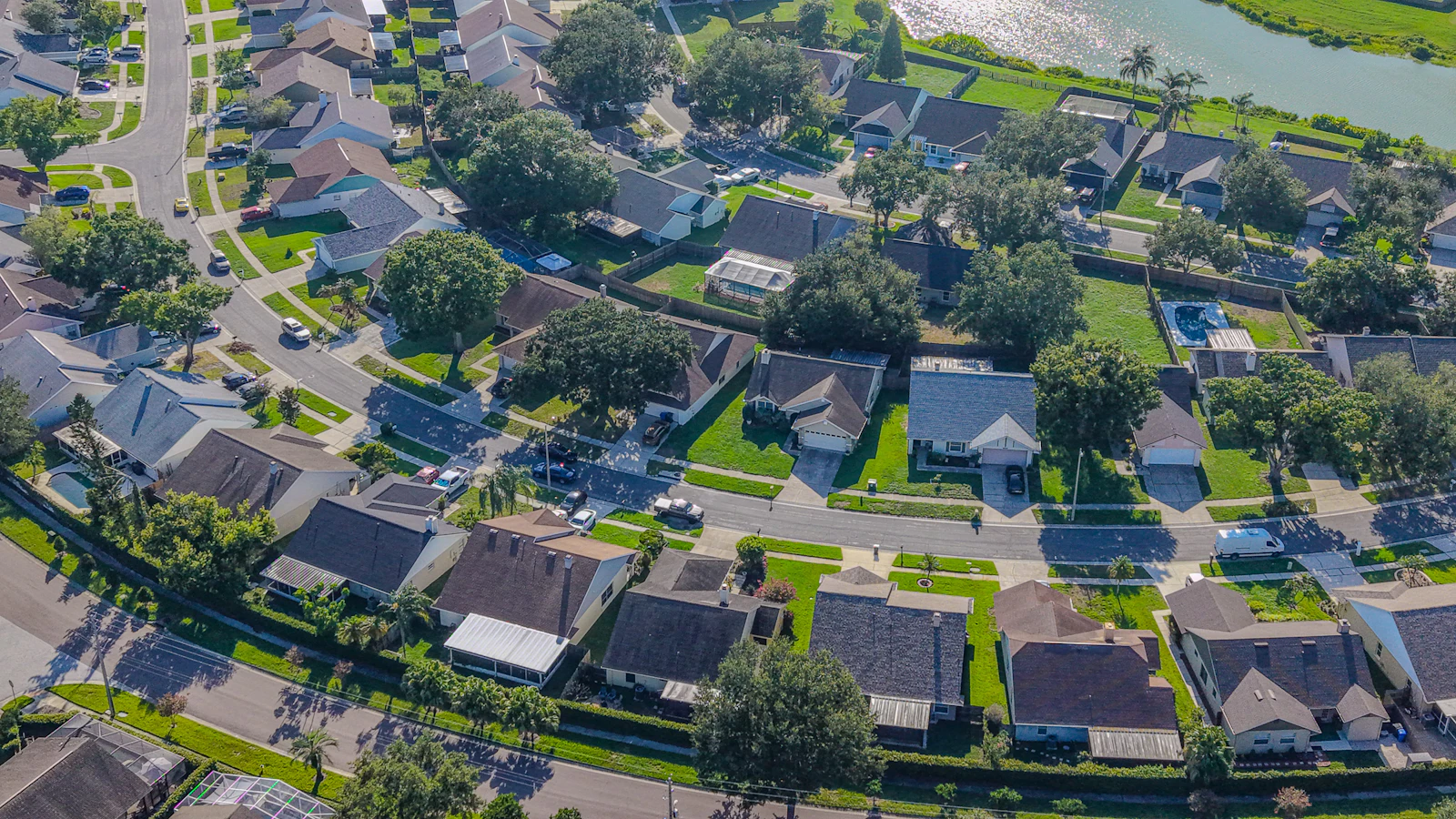 Vista aérea de bairro residencial com casas, ruas arborizadas, piscinas e um lago ao fundo.